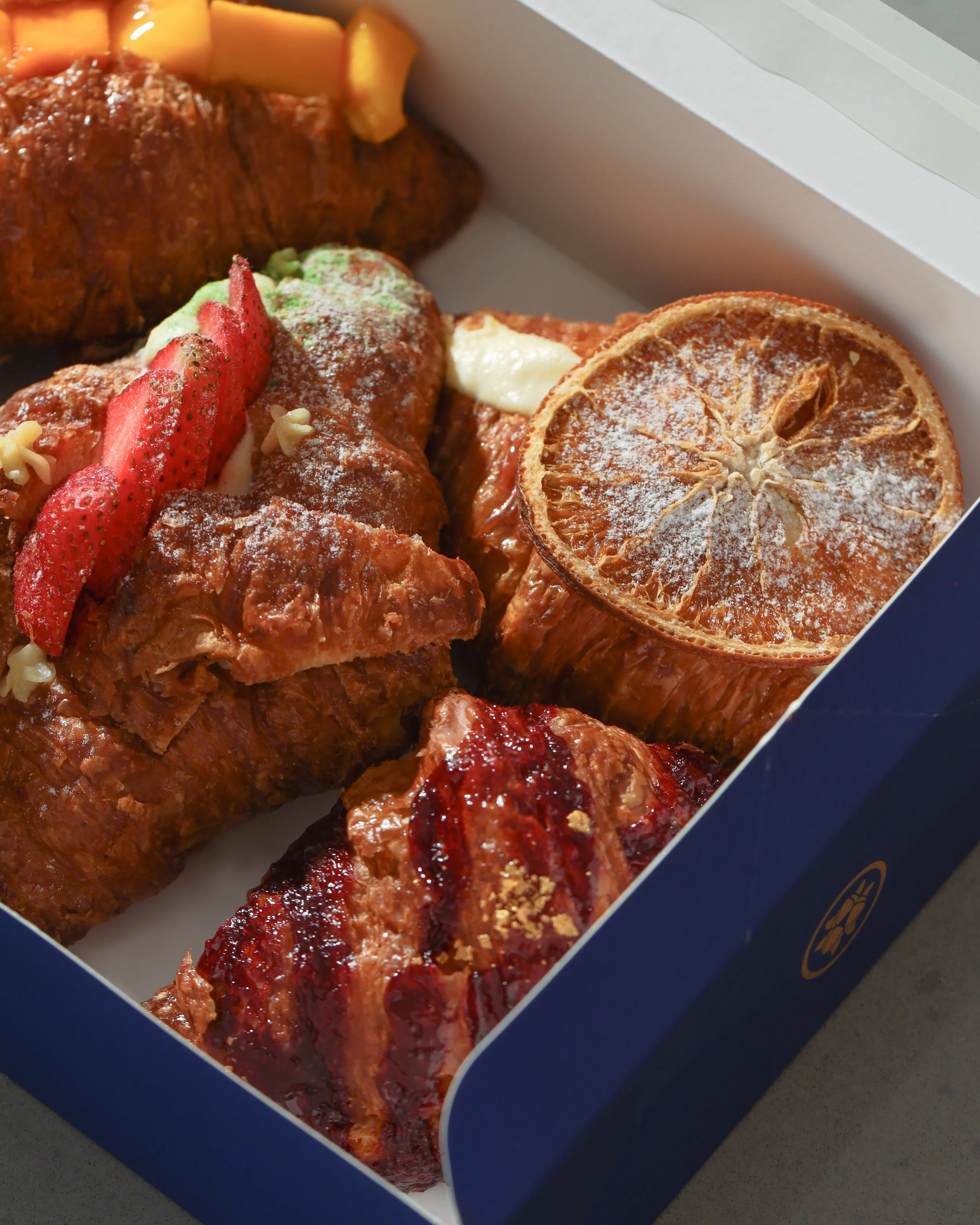 Close-up of a box of assorted baked goods, including flaky pastries topped with strawberries and powdered sugar, and slices of dried citrus fruit.