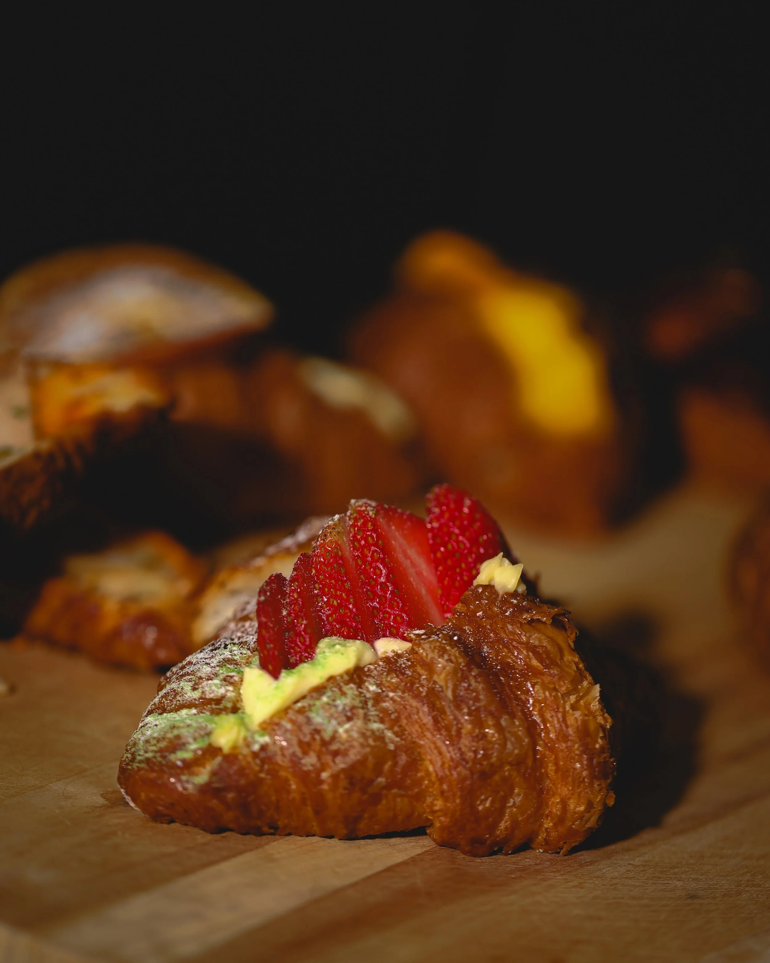 A croissant topped with sliced strawberries, small dollops of yellow cream, and a dusting of powdered sugar on a wooden surface.