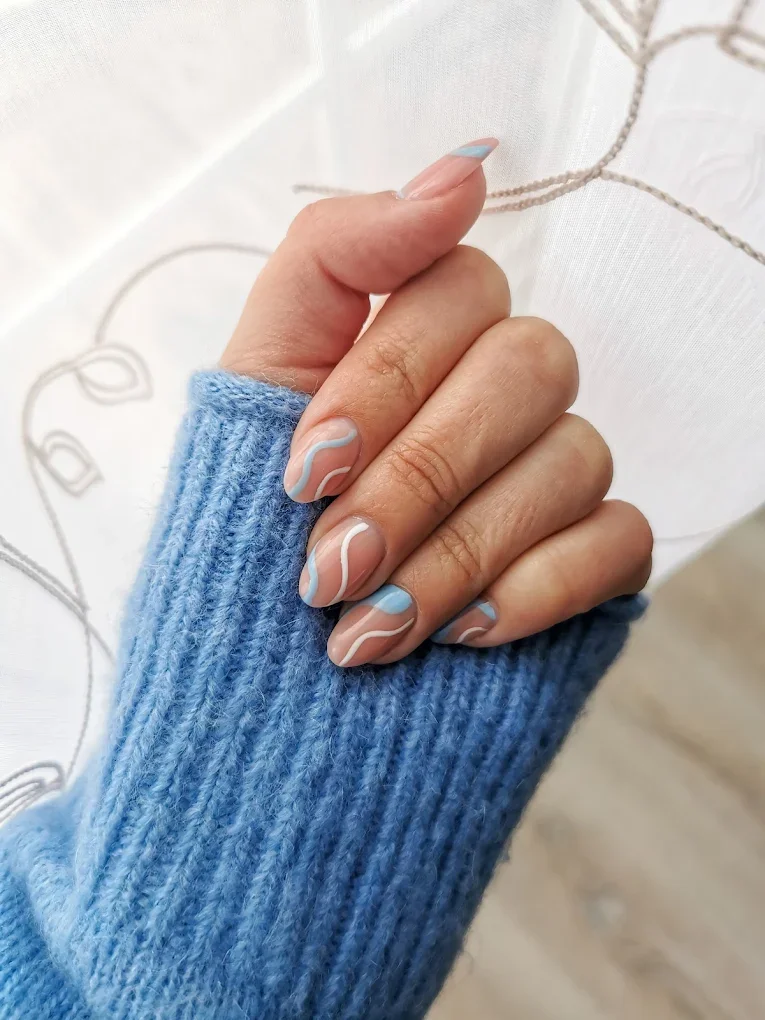 Hand mit blauer Strickjacke und French Maniküre mit blauen und weißen geschwungenen Linien auf den Nägeln, vor einem transparenten Vorhang.