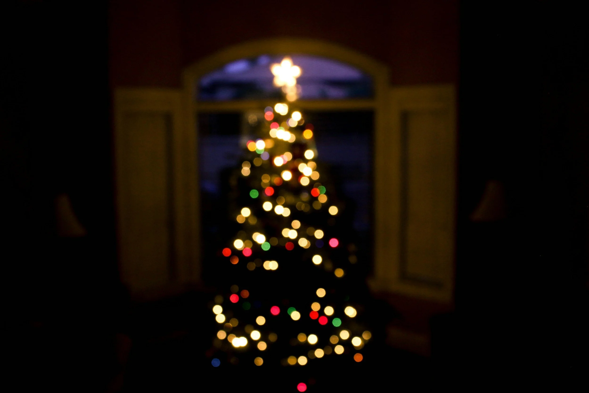 Blurred image of a decorated Christmas tree with colorful lights, viewed through a window at night.