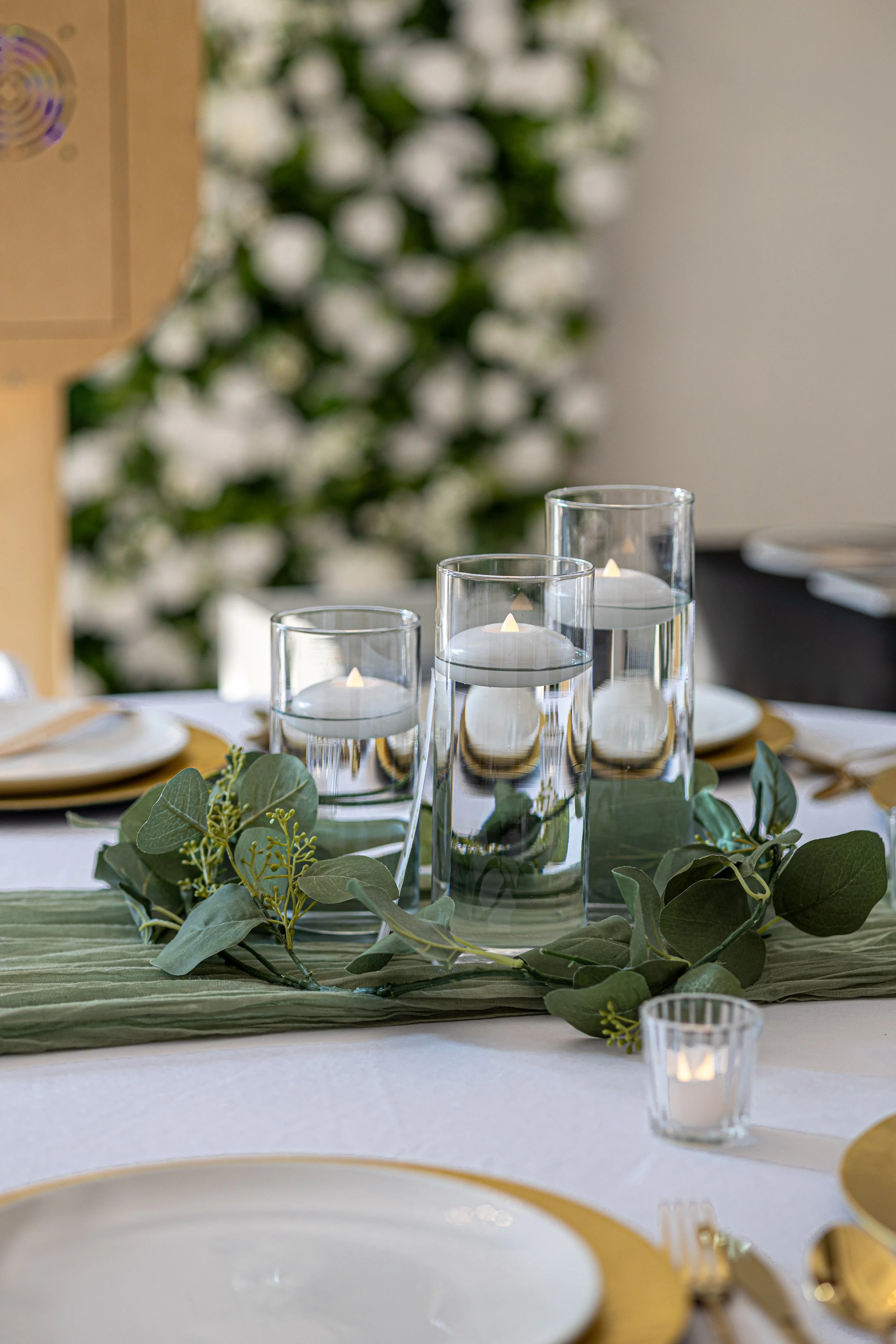 Table centerpiece with three glass vases containing floating candles, green foliage, and small candles in votive holders on a white tablecloth. Wedding photographer