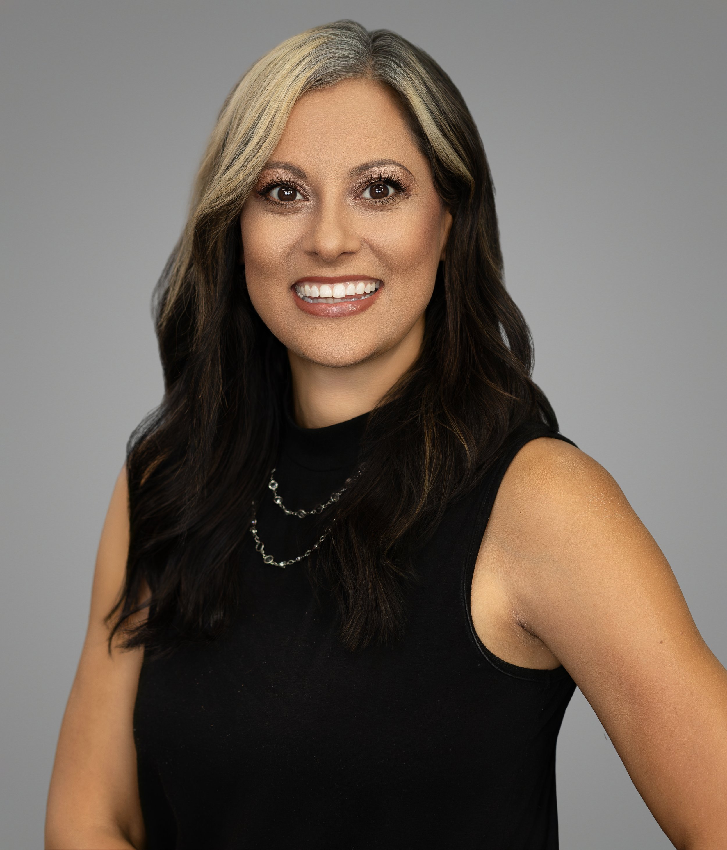 Close-up photo of a smiling woman with long dark hair, wearing a black sleeveless top and a silver necklace, against a plain gray background.