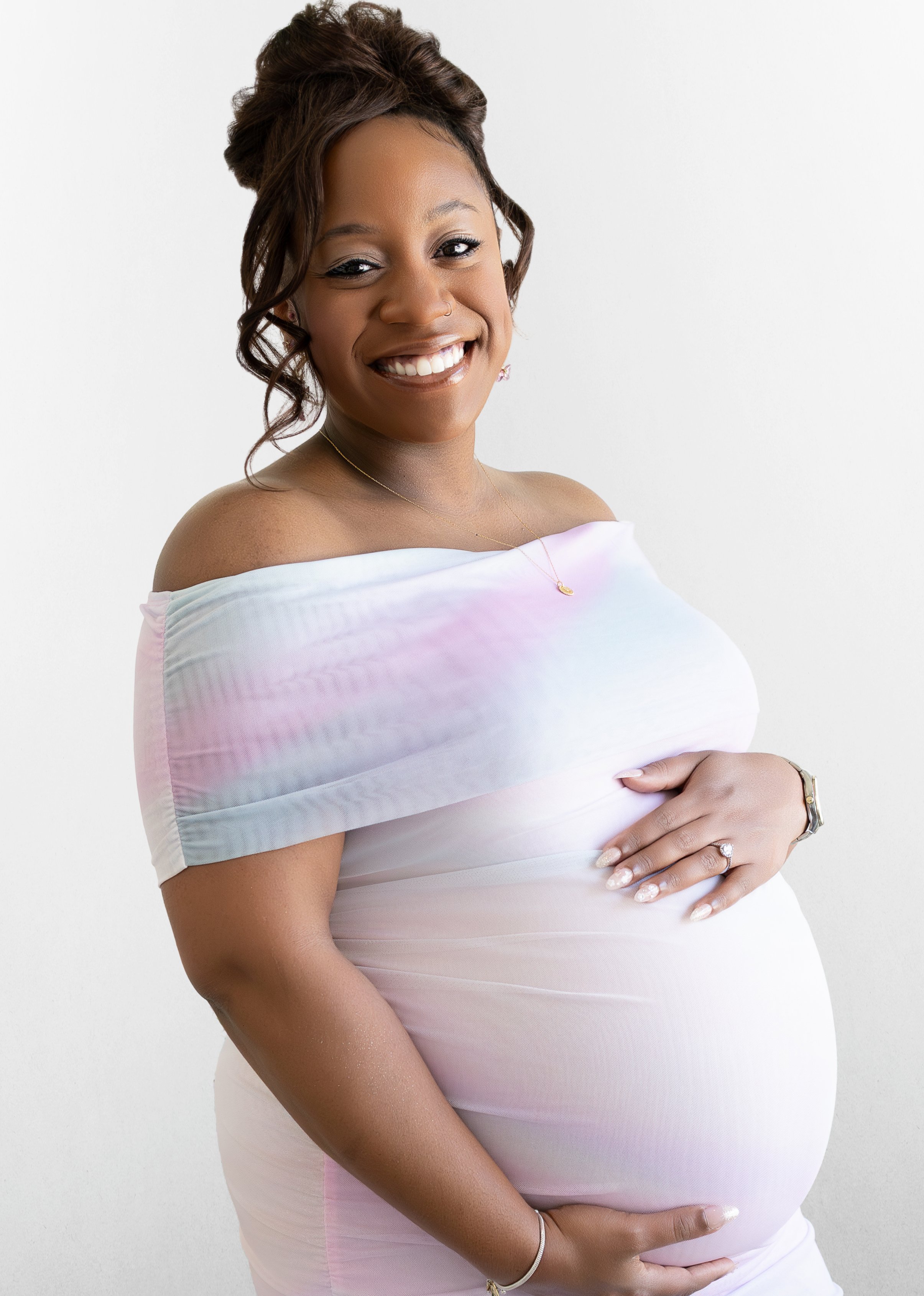 A smiling pregnant woman with dark hair styled in curls, wearing a pastel off-shoulder top, gold necklace, and jewelry, gently cradling her baby bump against a plain white background.