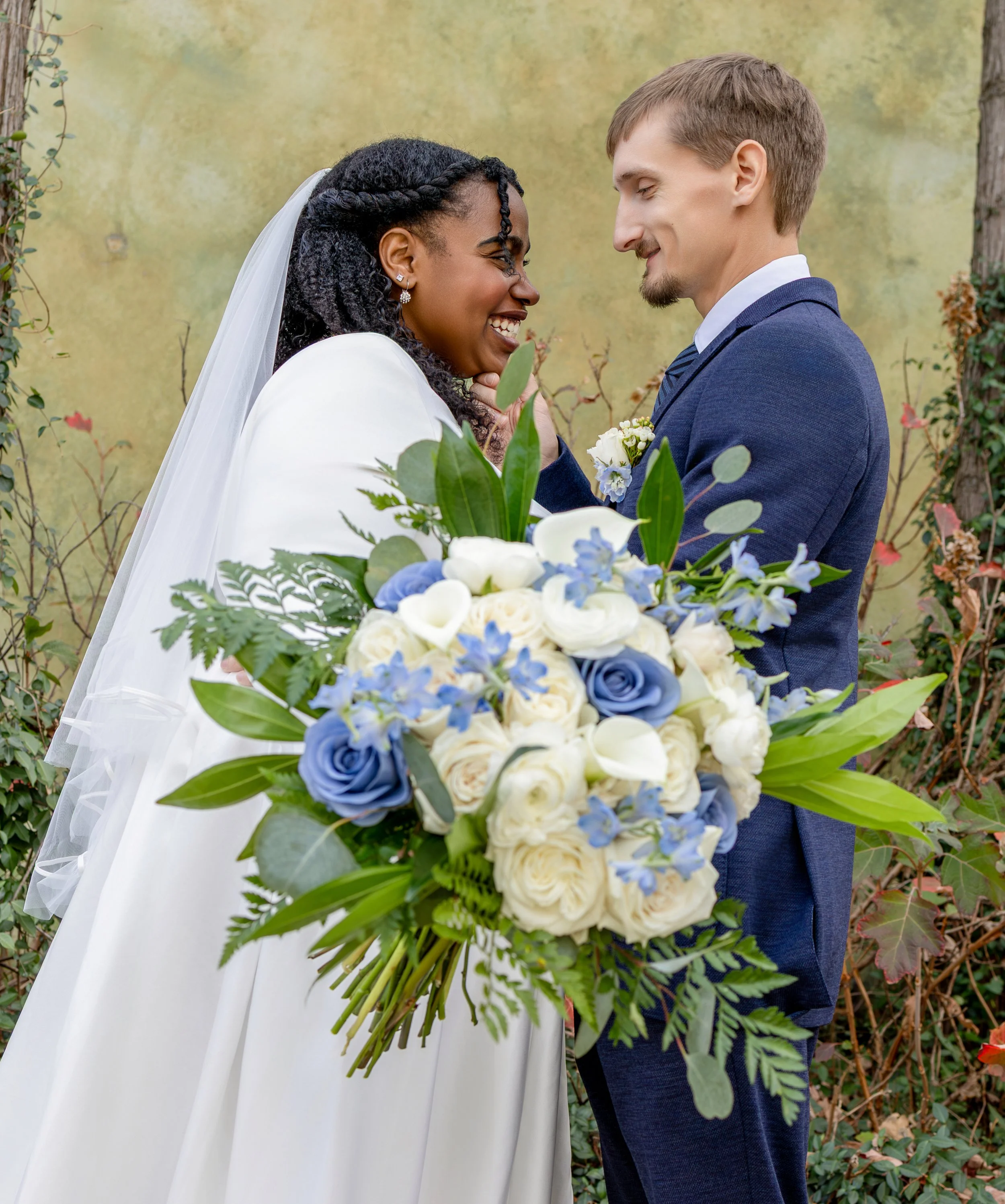 A bride and groom share a joyful moment outdoors, with the bride holding a large bouquet of white and blue flowers, and they stand close, smiling at each other. wedding photography dfw photography