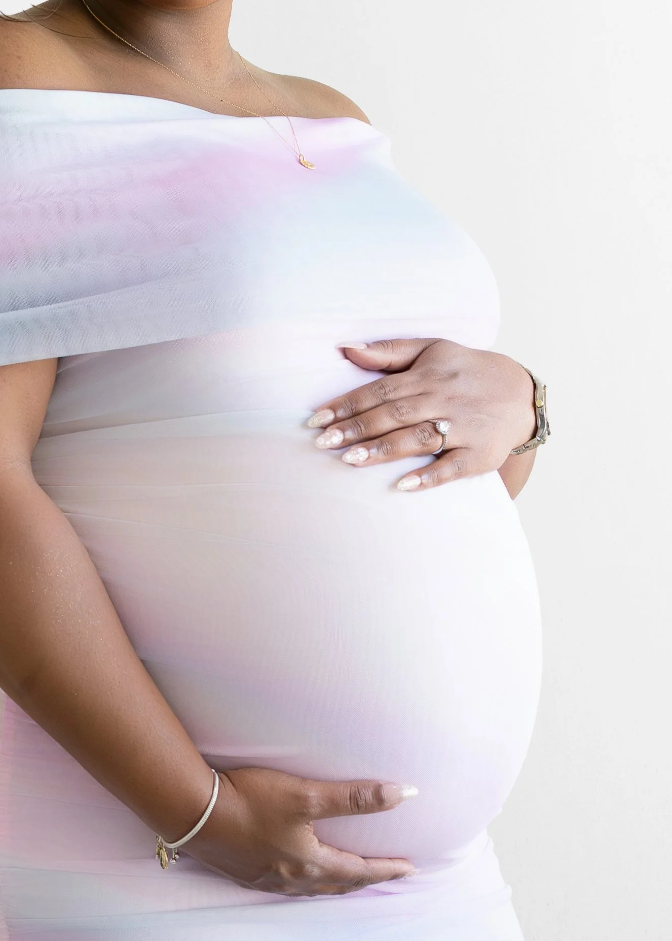 A pregnant woman wearing a white off-the-shoulder dress, holding her belly with one hand and gently touching her stomach with the other, against a plain white background.
