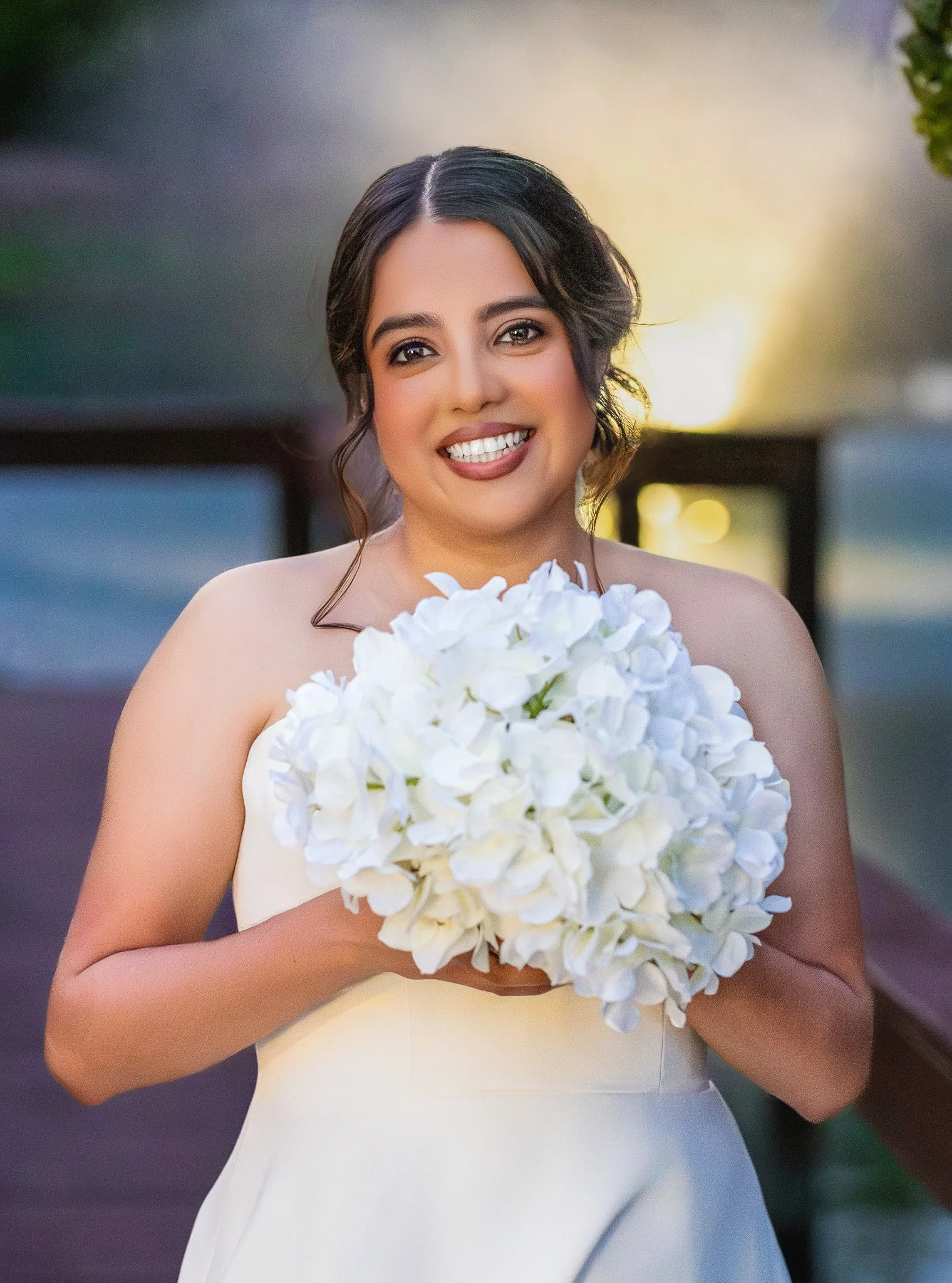 A bride in a white dress holding a bouquet of white hydrangeas, smiling outdoors during sunset. wedding photographer