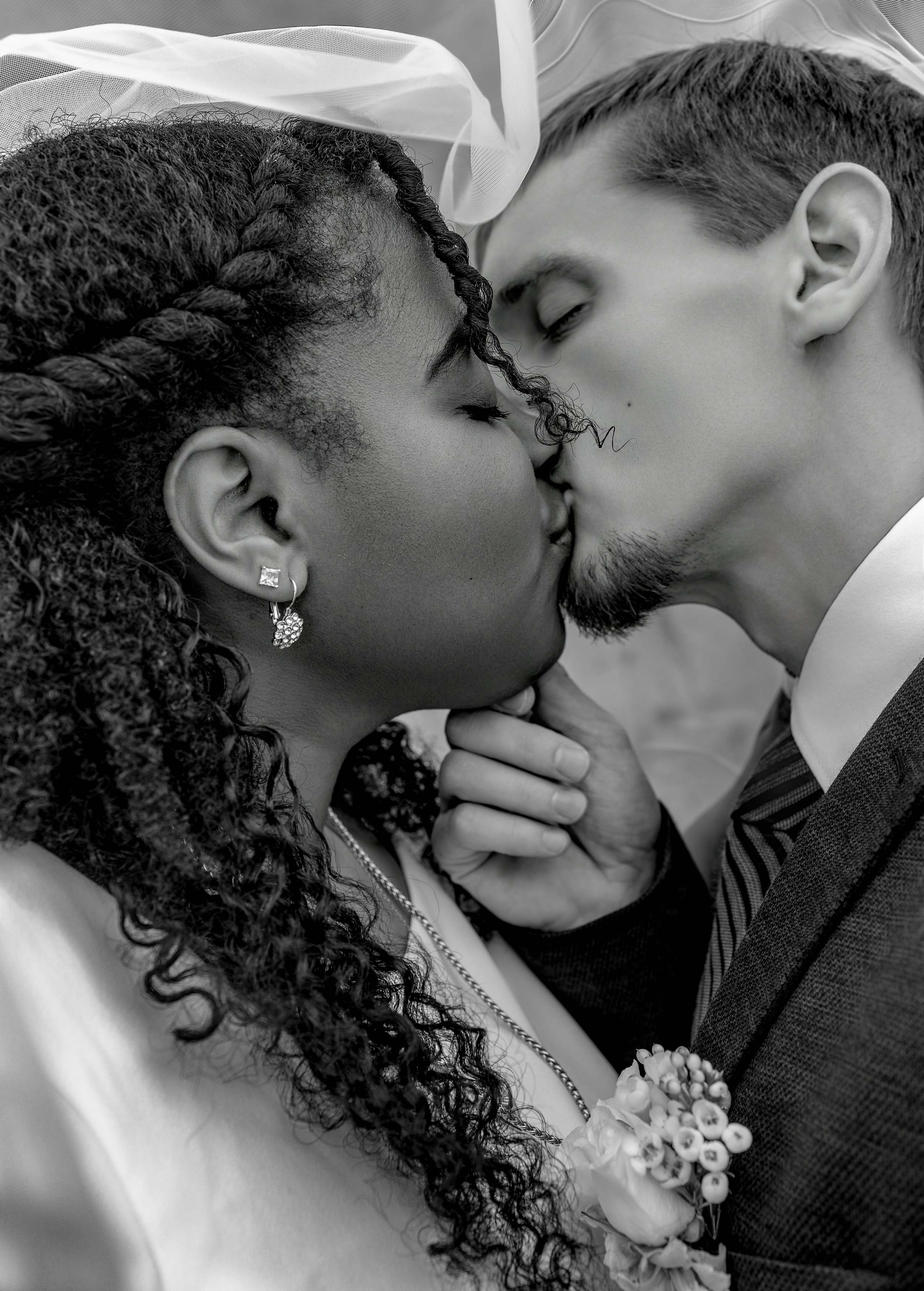 A black and white photograph of a couple kissing, with their eyes closed. The woman has curly hair styled in braids and is wearing earrings, a necklace, and a white dress. The man has short hair, a beard, and is dressed in a suit with a striped tie, holding a bouquet of flowers.