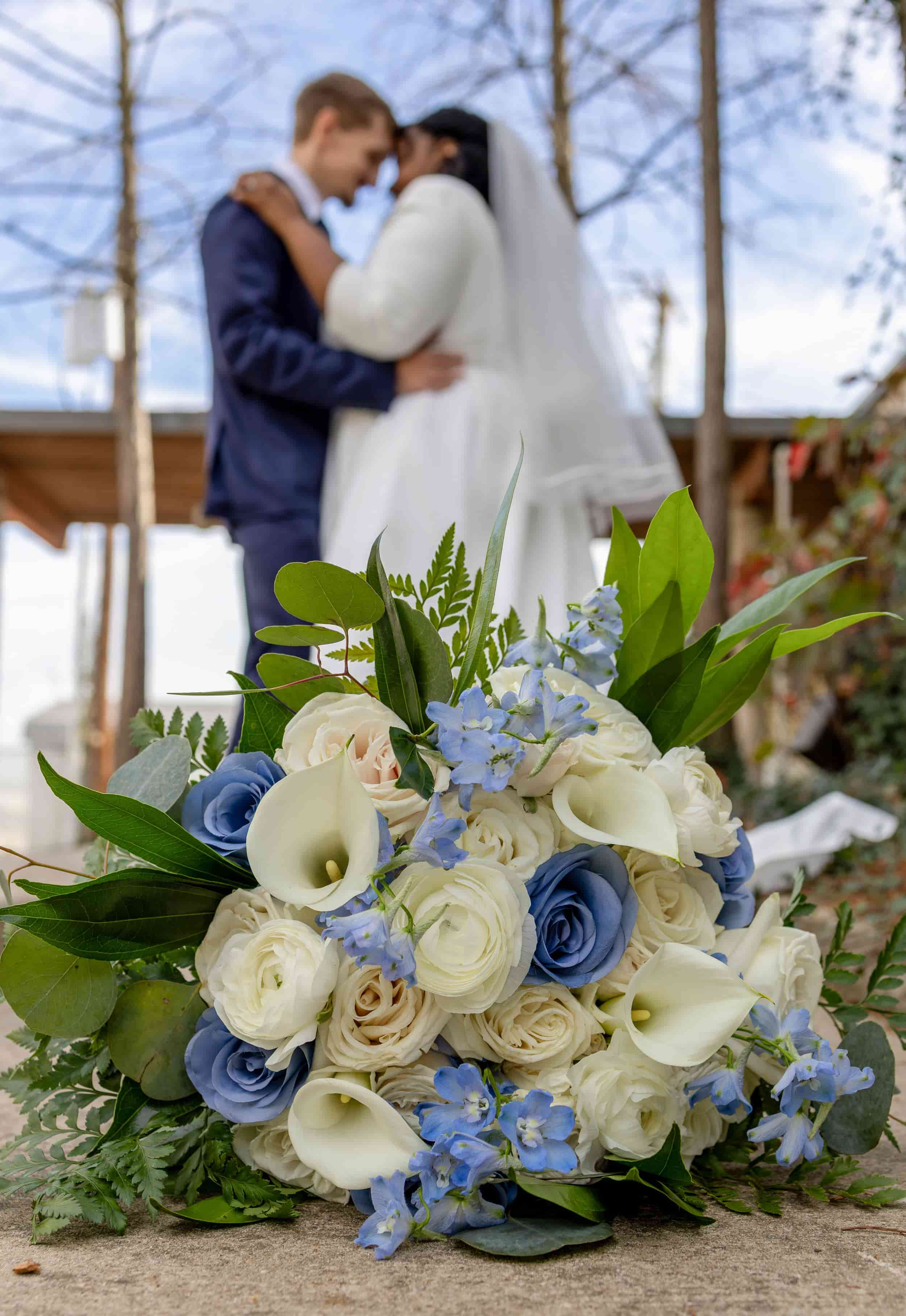 A wedding bouquet of white and blue roses, calla lilies, and assorted greenery, placed on a surface with a blurred background of a couple in wedding attire embracing. wedding photography