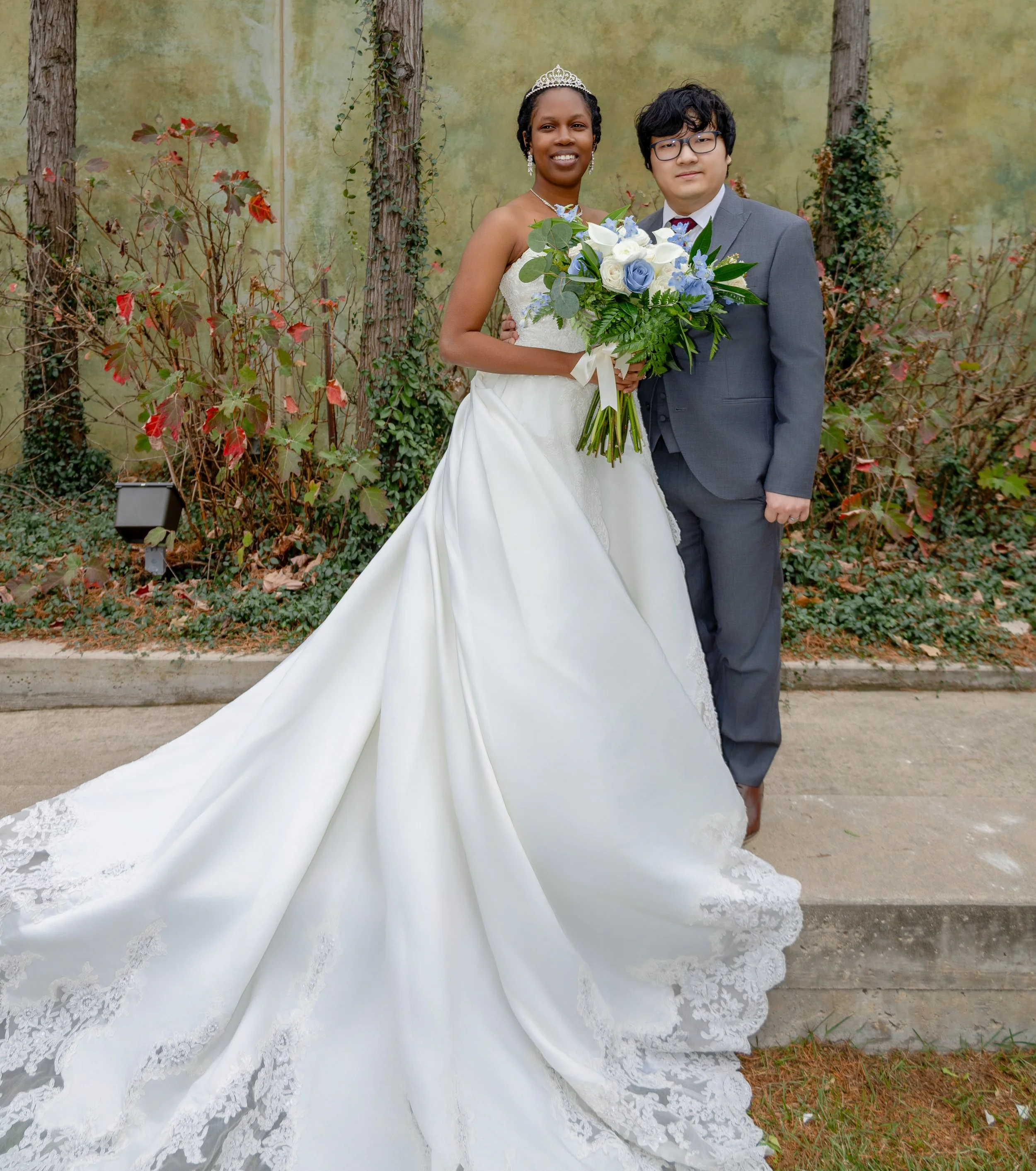 A bride in a white wedding dress holding a bouquet standing next to a groom in a gray suit. They are outdoors with a green wall and plants in the background. wedding photography dfw photography