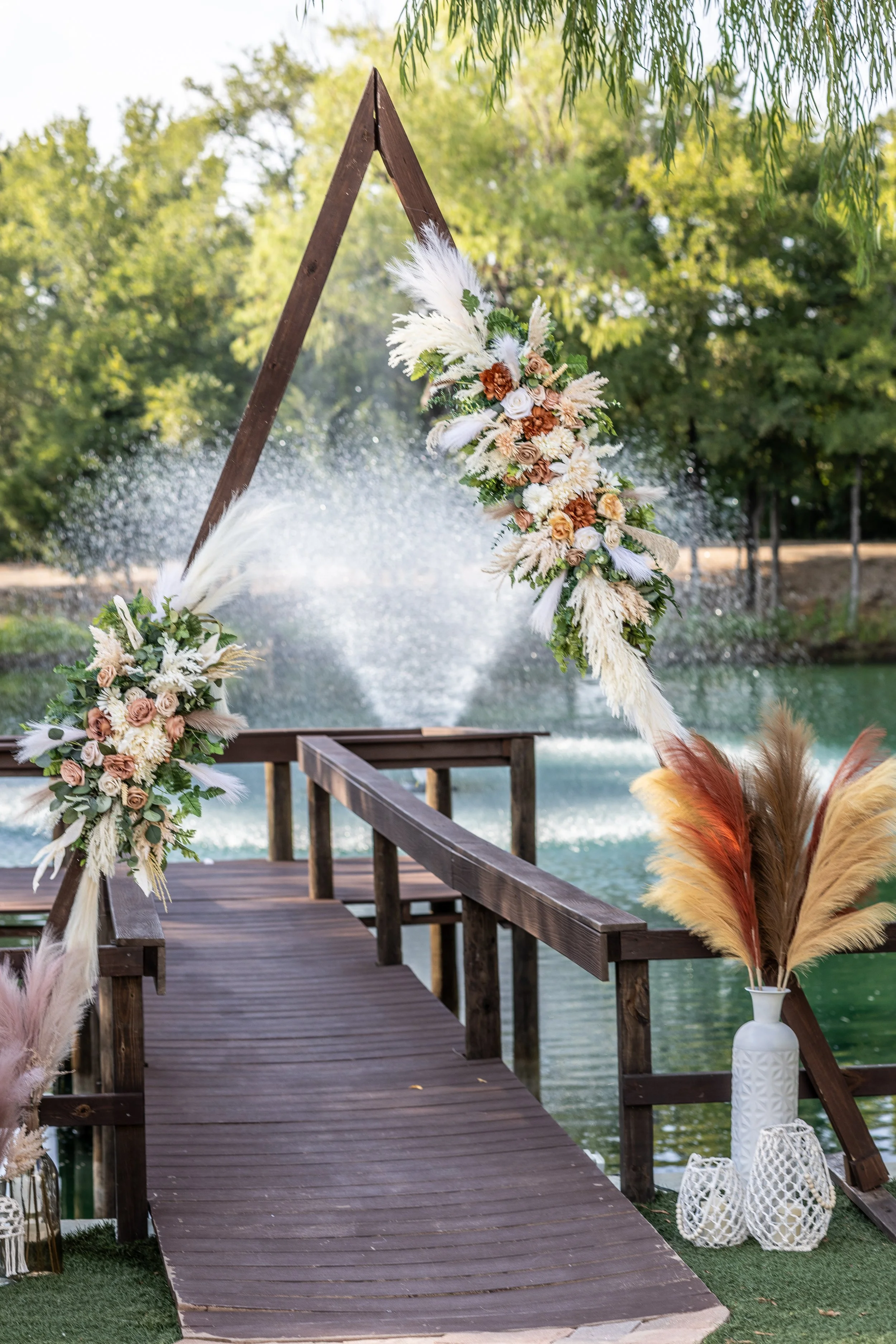 Outdoor wedding altar decorated with flowers and pampas grass, set on a wooden dock by a lake with trees in the background. wedding photographer