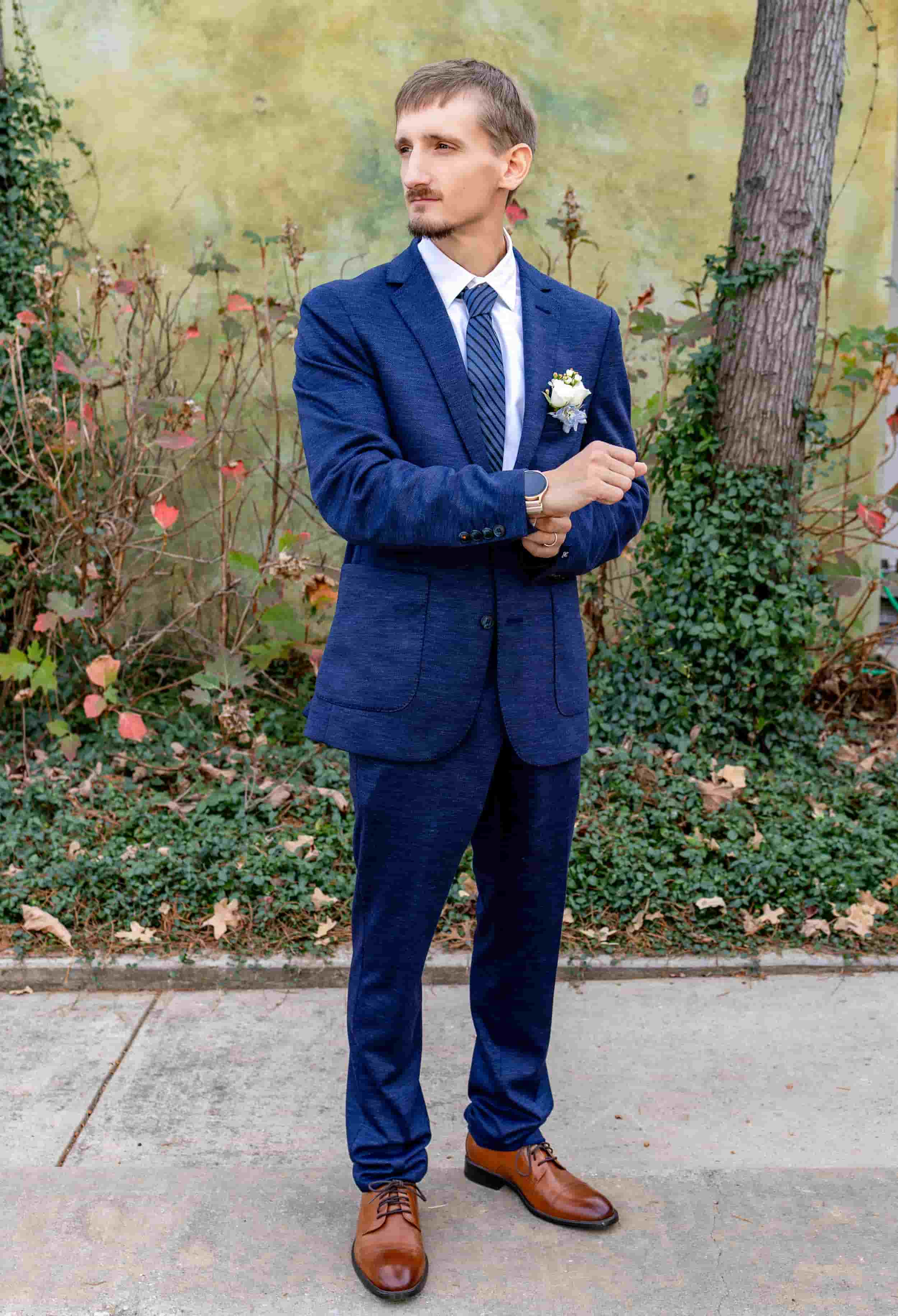 Man in a blue suit with a white shirt and striped tie, standing outdoors near a tree, with a white boutonniere on his lapel, looking to the side. wedding photography