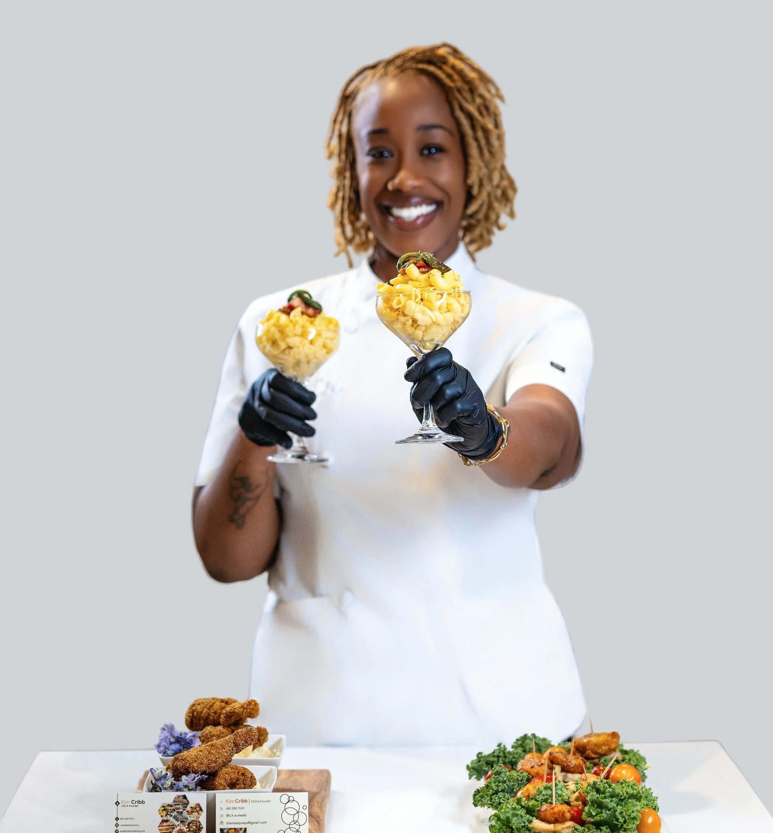 Woman in a white chef's coat and black gloves holding two dessert glasses filled with pasta salad, smiling. DFW Photographer - Branding Photography
