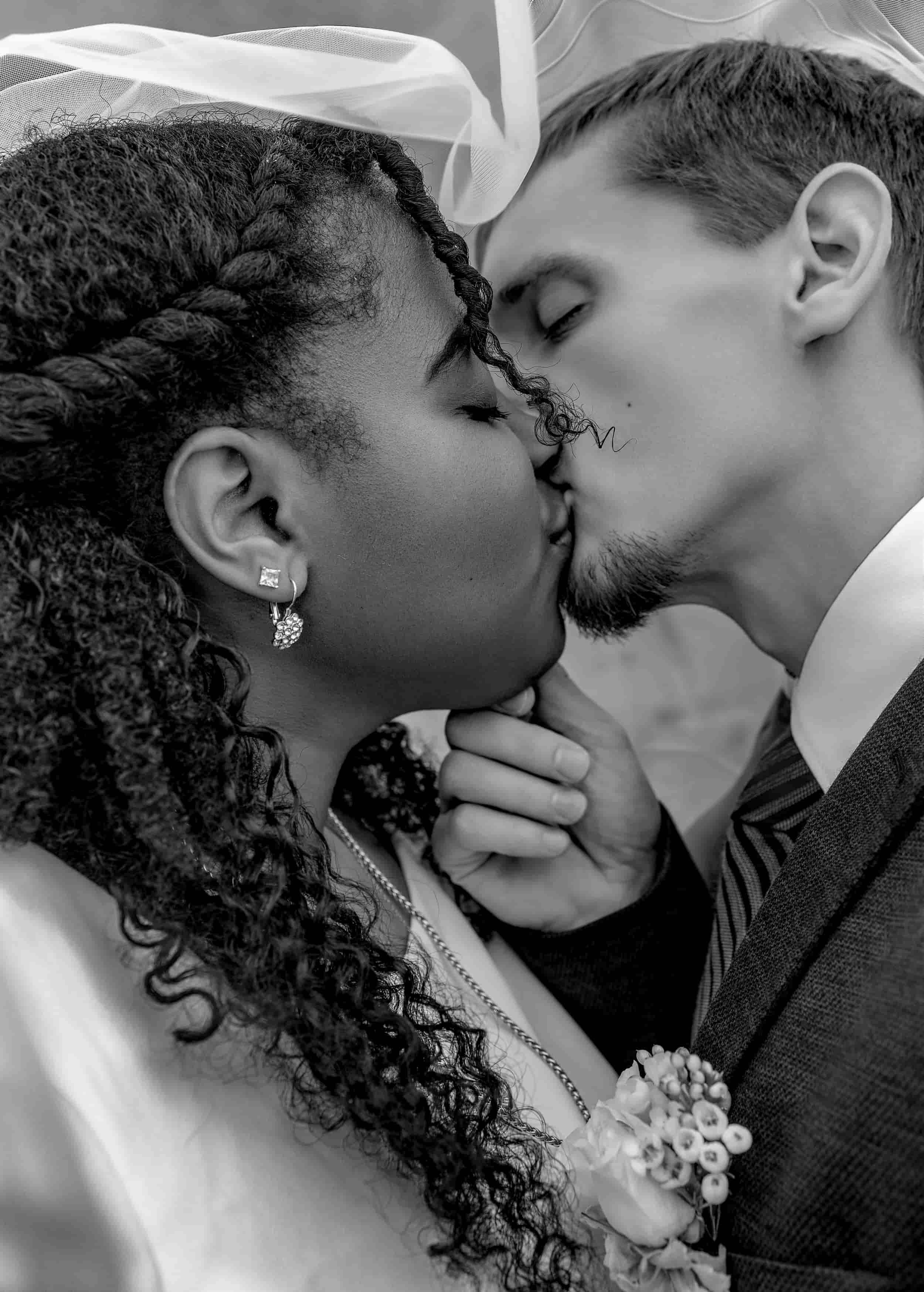 A black-and-white photograph of a couple kissing, with the woman wearing earrings and a bouquet pinned to her dress, and the man in a suit with a striped tie. wedding photography