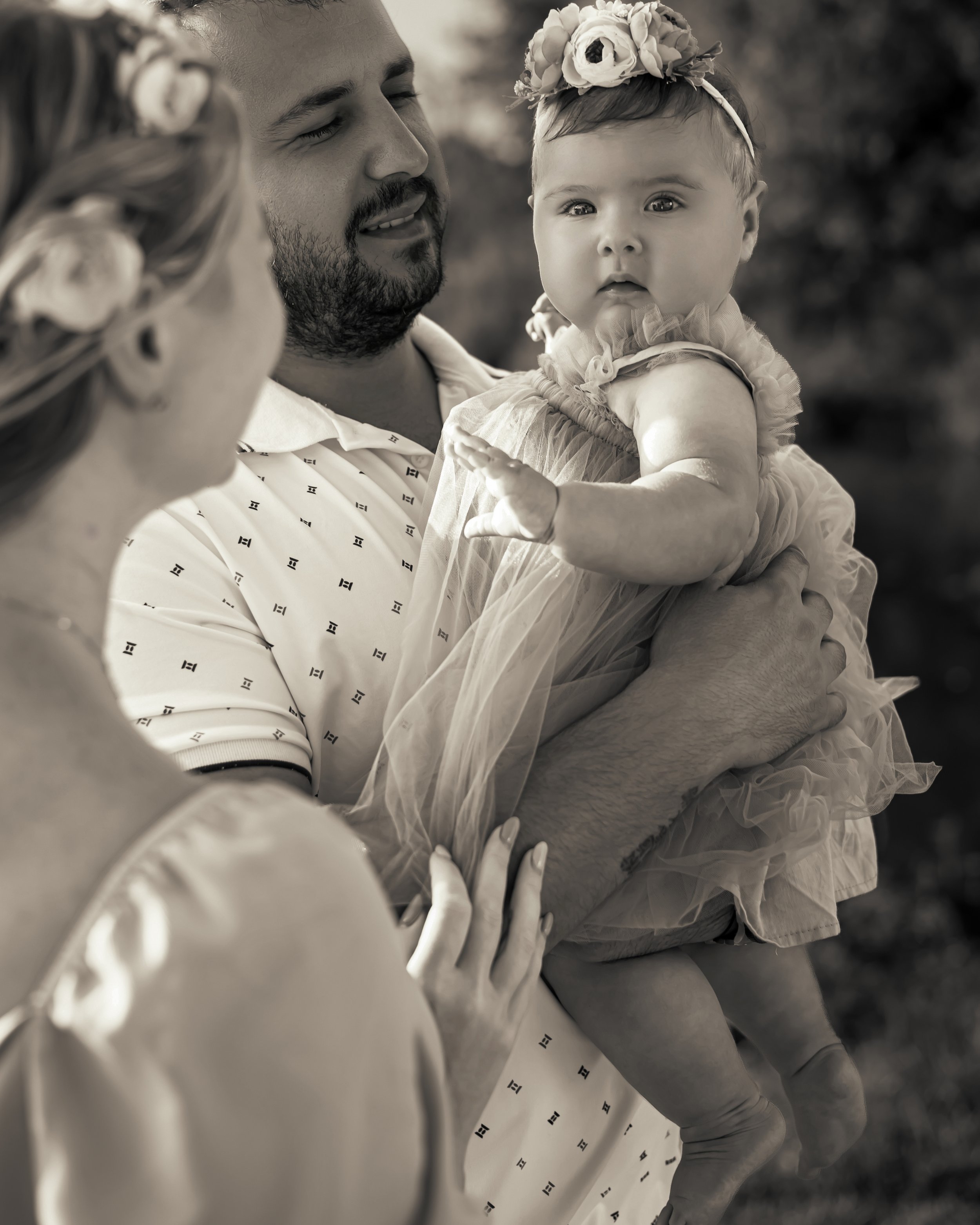 A man holds a baby girl wearing a flower headband and a frilly dress, while a woman with curly hair, seen from behind, is partially visible in the foreground. The scene appears to be outdoors, possibly during a family gathering or celebration.