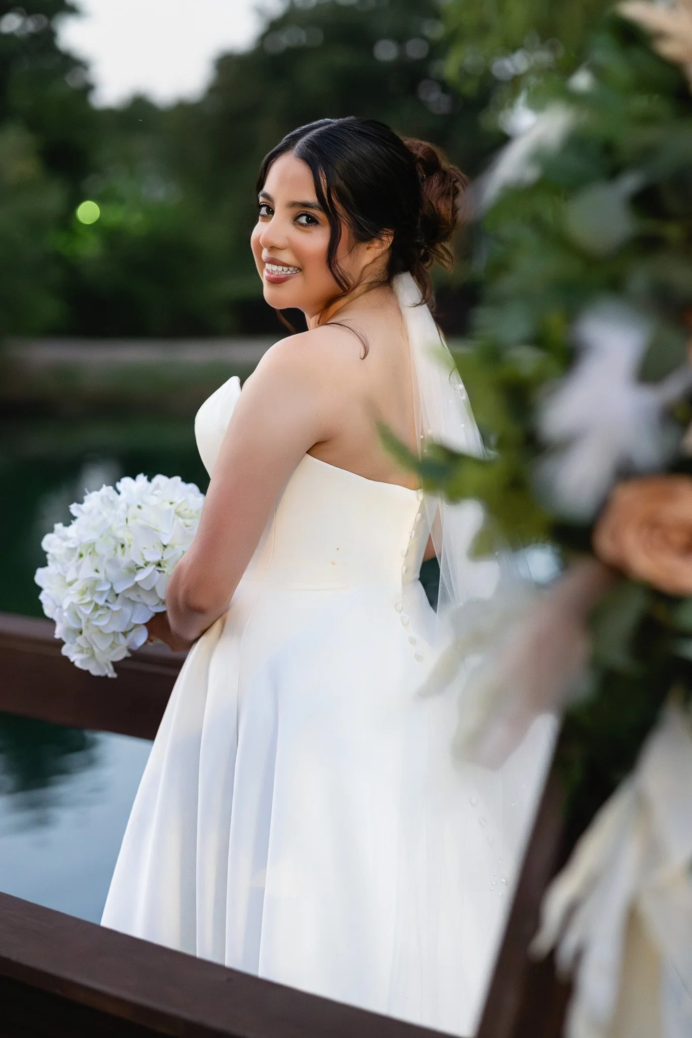 A bride in a white wedding dress holding a bouquet of white flowers, smiling, outdoors near water with greenery in the background. wedding photographer