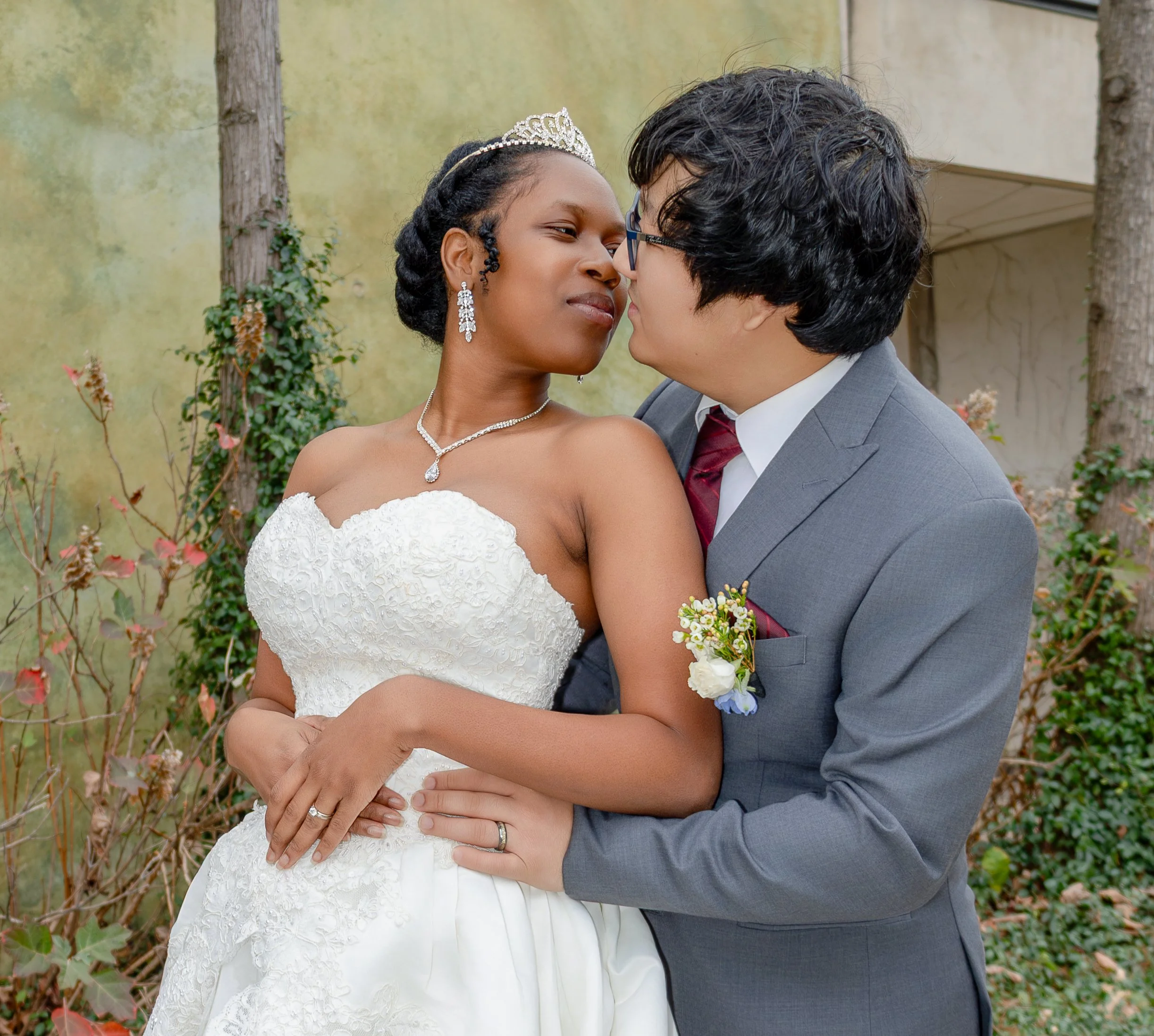 A bride and groom share an intimate moment outdoors, with the bride wearing a strapless lace wedding gown, a tiara, and jewelry, and the groom in a gray suit with a boutonniere and glasses, surrounded by greenery. wedding photography dfw photography