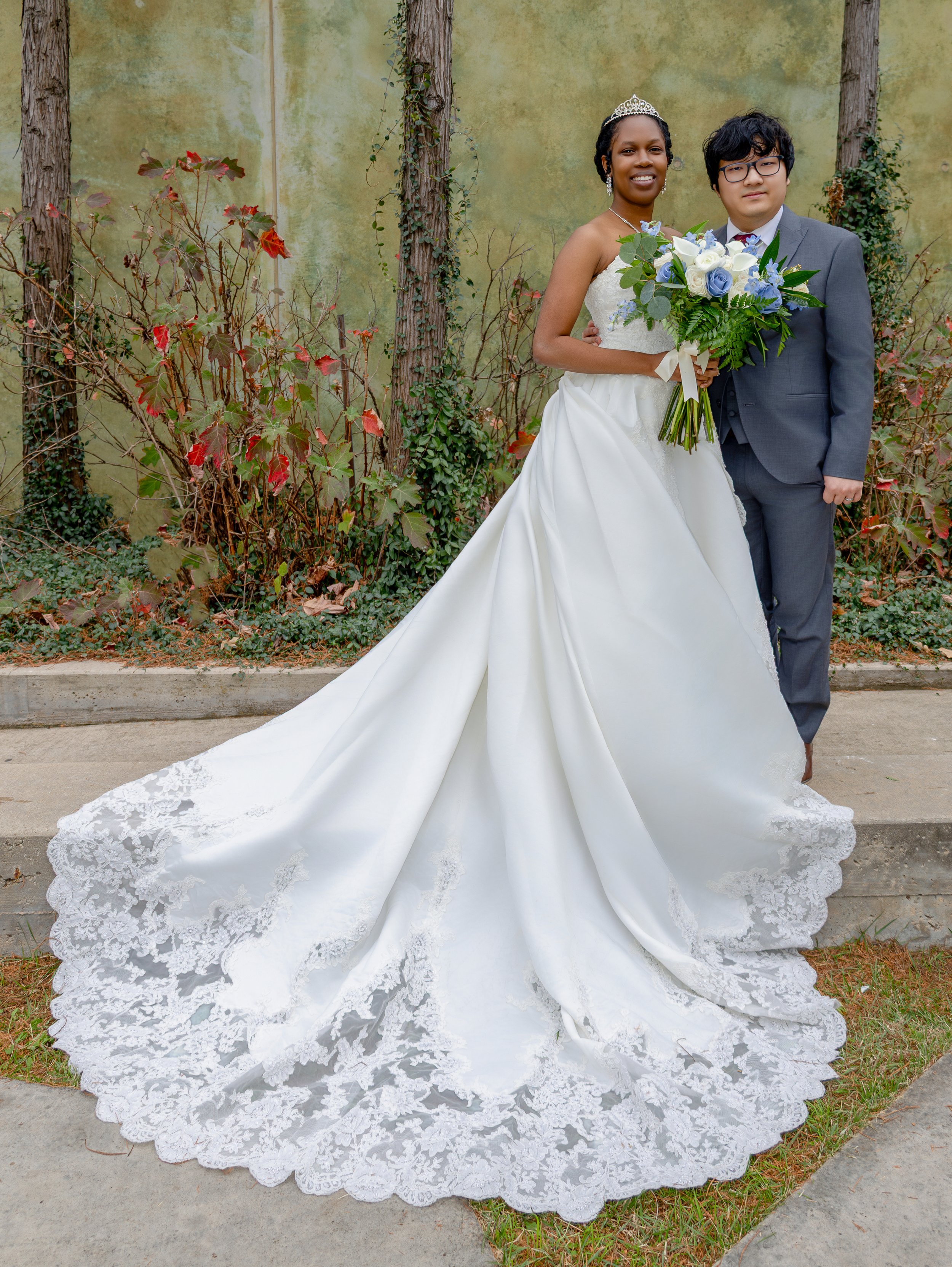 A bride in a white wedding gown with lace detailing and a groom in a gray suit, standing outdoors with a green wall and foliage background, holding a bouquet of white and blue flowers. wedding photography dfw photography