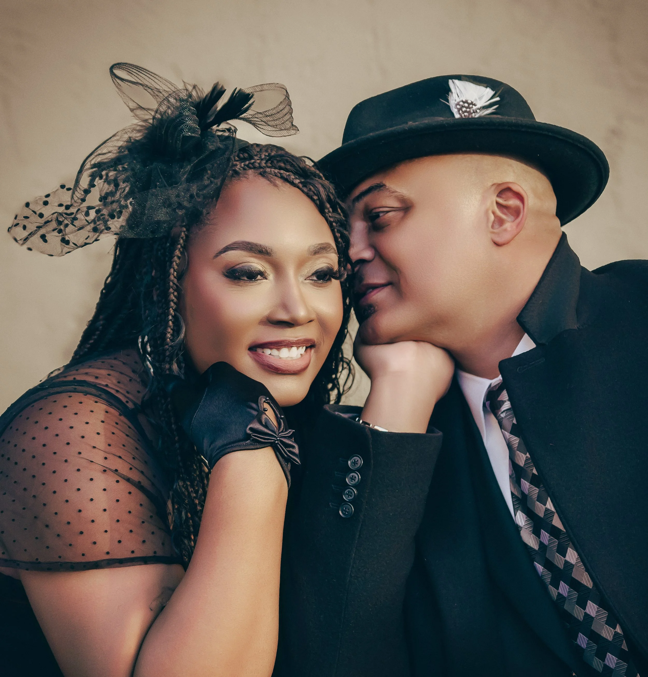 Close-up of a couple dressed in formal attire, with the woman wearing a tulle hat and the man wearing a black fedora with a feather decoration, leaning in close to each other.
