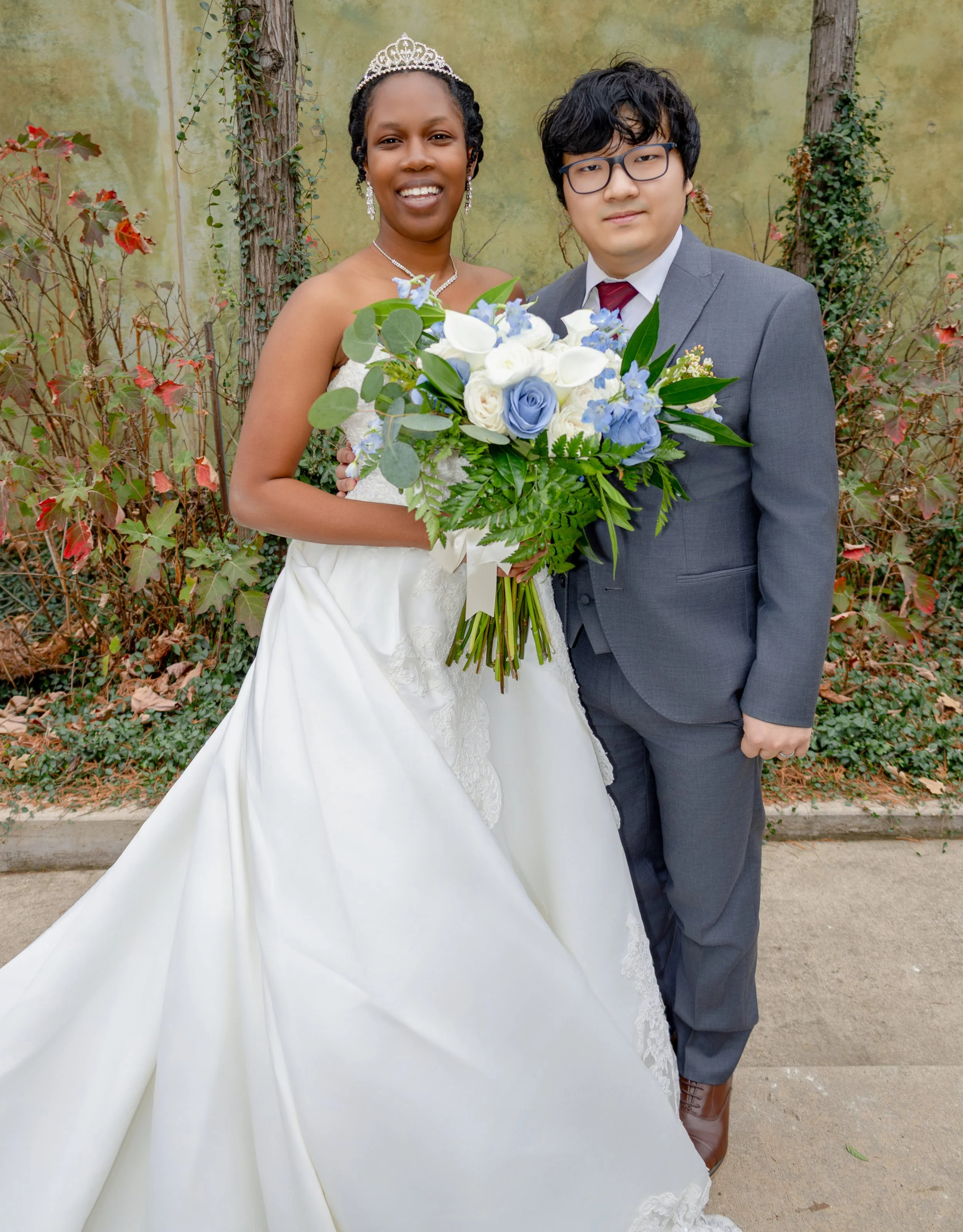 A bride and groom standing outdoors with a floral backdrop. The bride is wearing a white wedding gown with lace details, a tiara, and jewelry, holding a large bouquet of white and blue flowers. The groom is in a gray suit with glasses and a white shi