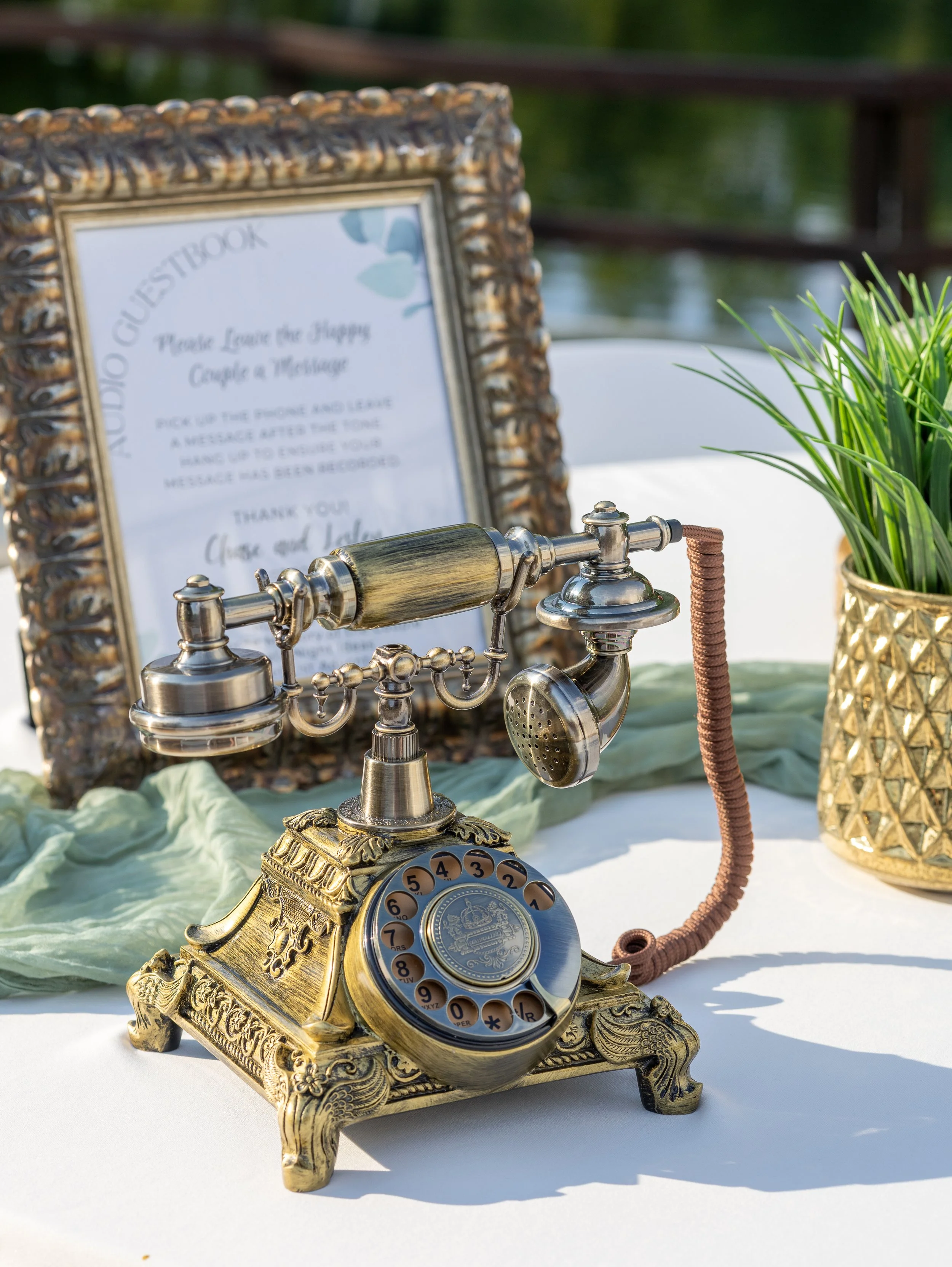Decorative vintage rotary telephone with gold and silver finish on a white table, beside a framed sign with instructions, and a gold vase with green plants at an outdoor event. wedding photographer