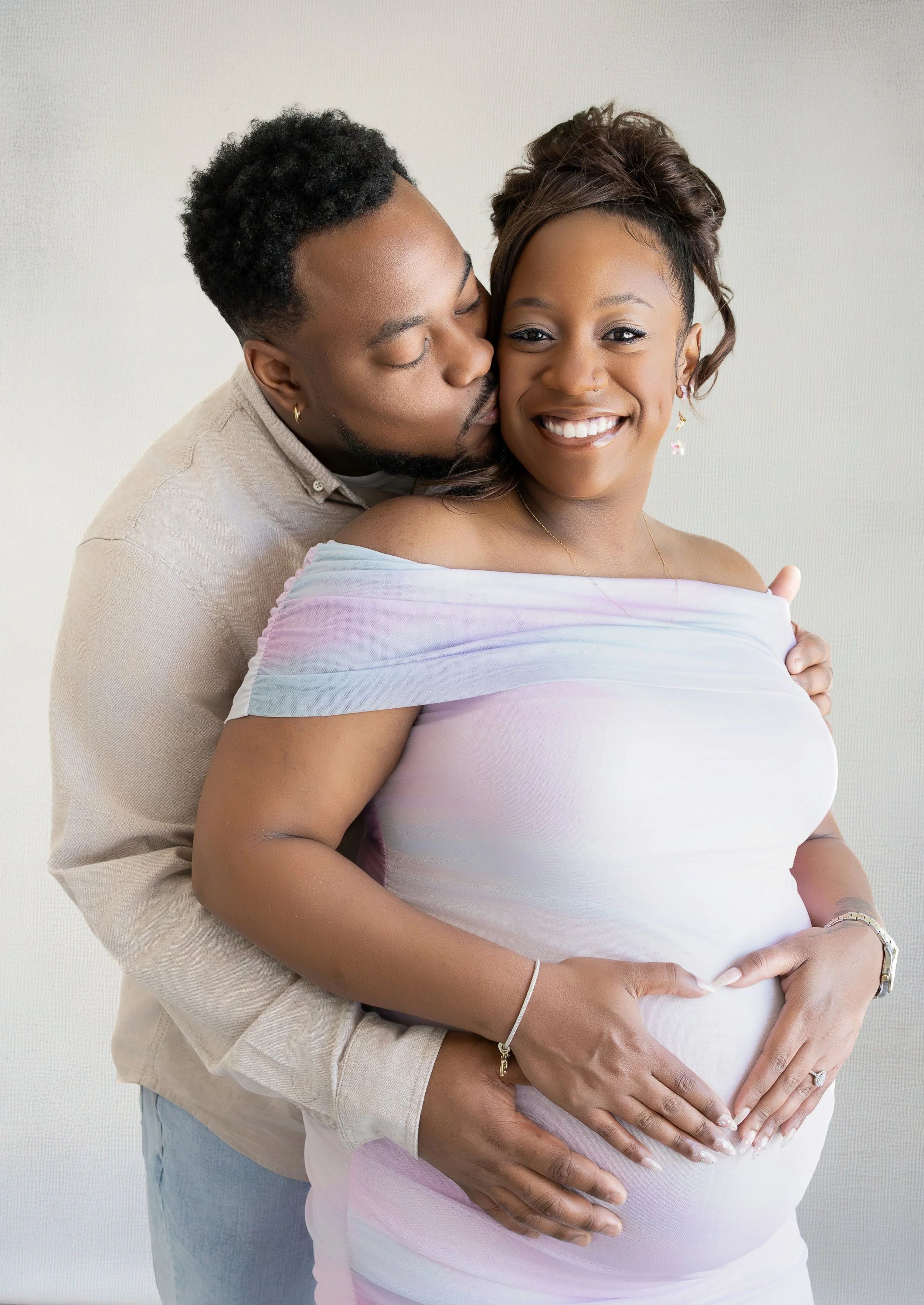 A man and a pregnant woman are standing close together, with the man kissing the woman on the cheek. The woman is smiling and forming a heart shape with her hands over her pregnant belly. They are indoors against a plain light-colored wall.