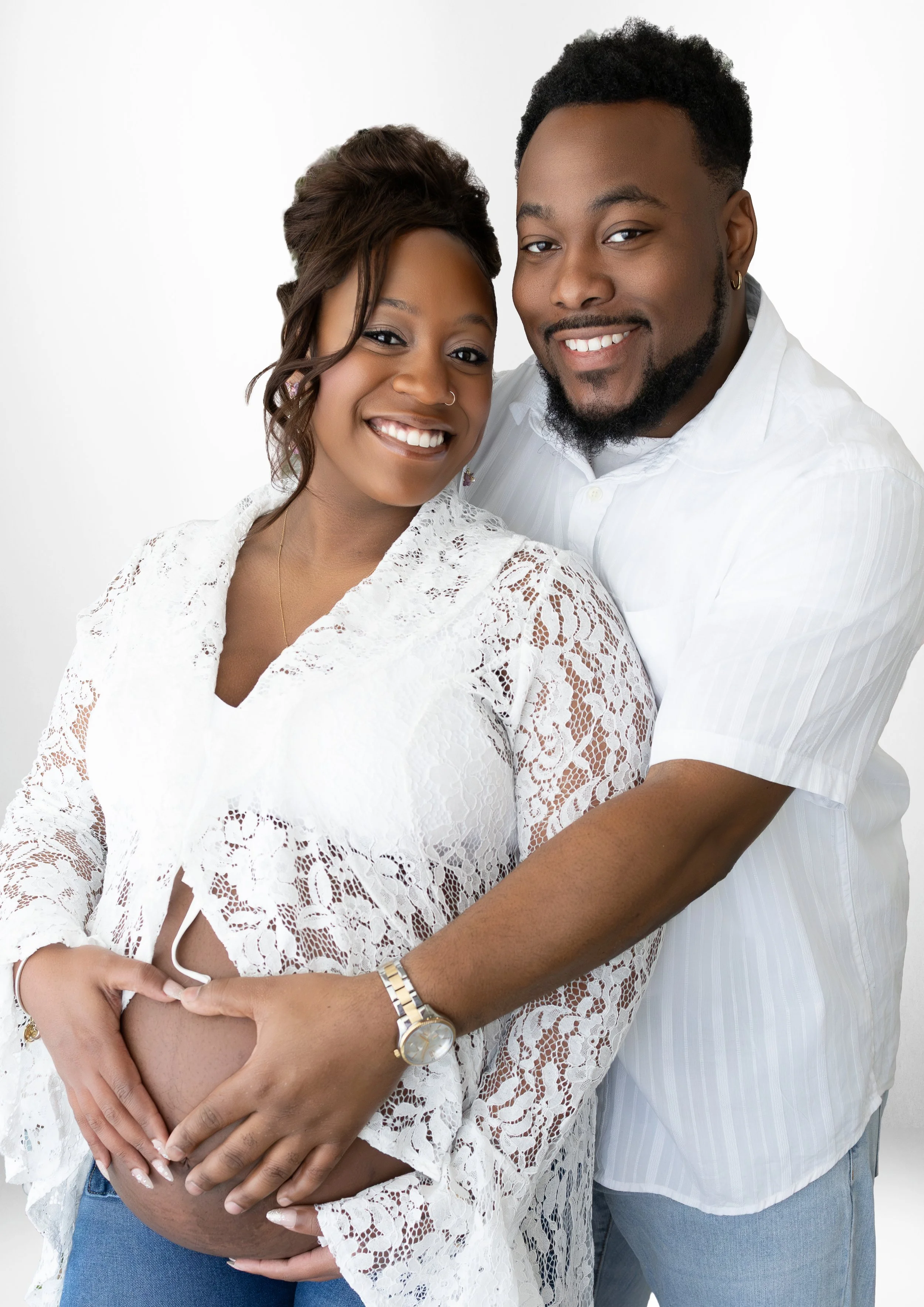 A pregnant woman in a white lace top and jeans is hugging her belly, while a man in a striped white shirt and blue jeans embraces her from behind, both smiling at the camera against a plain white background.