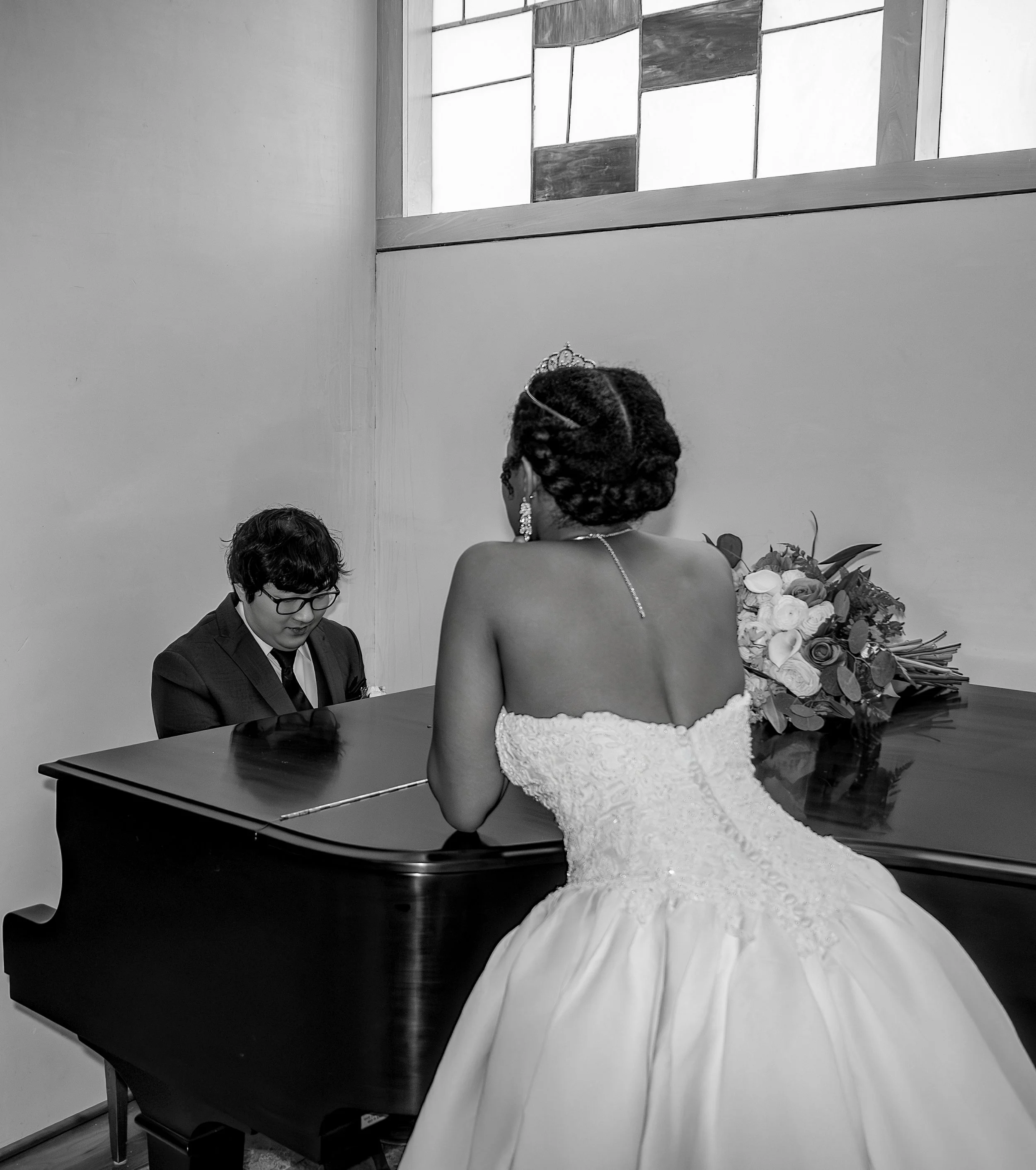 A bride in a strapless wedding gown with intricate embroidery gazes at a man in a suit playing piano. The bride wears a tiara and earrings, with a bouquet of flowers on the piano. wedding photography dfw photography