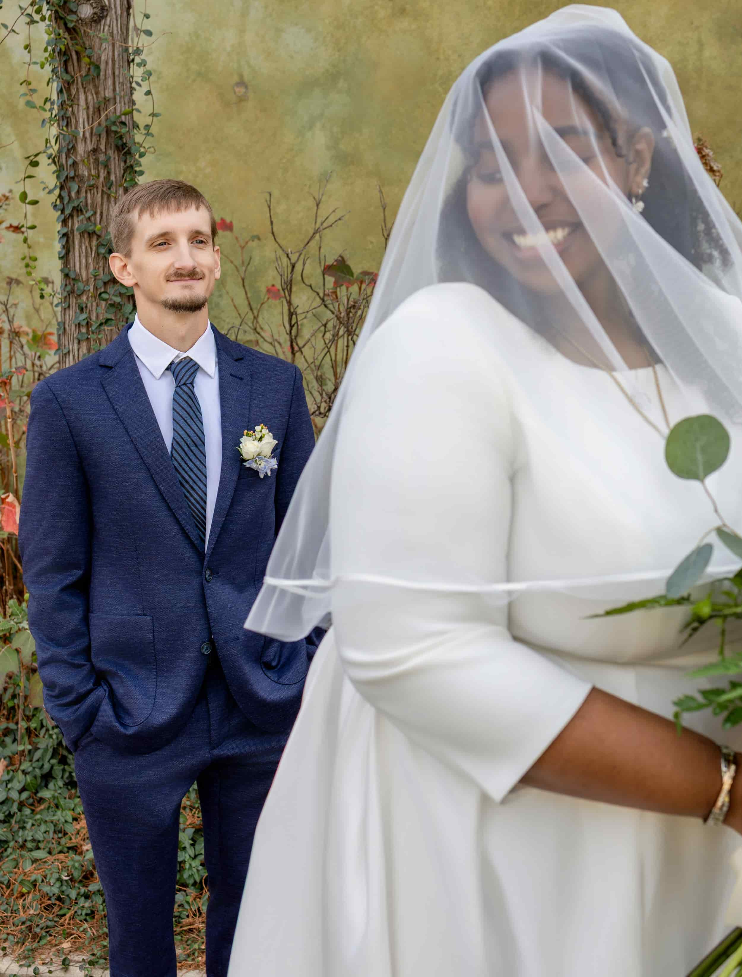 A bride in a white wedding dress with a veil, smiling, holding a bouquet, and a groom in a navy suit with a striped tie and boutonniere, standing outside against a natural background with greenery and a tree. wedding photography
