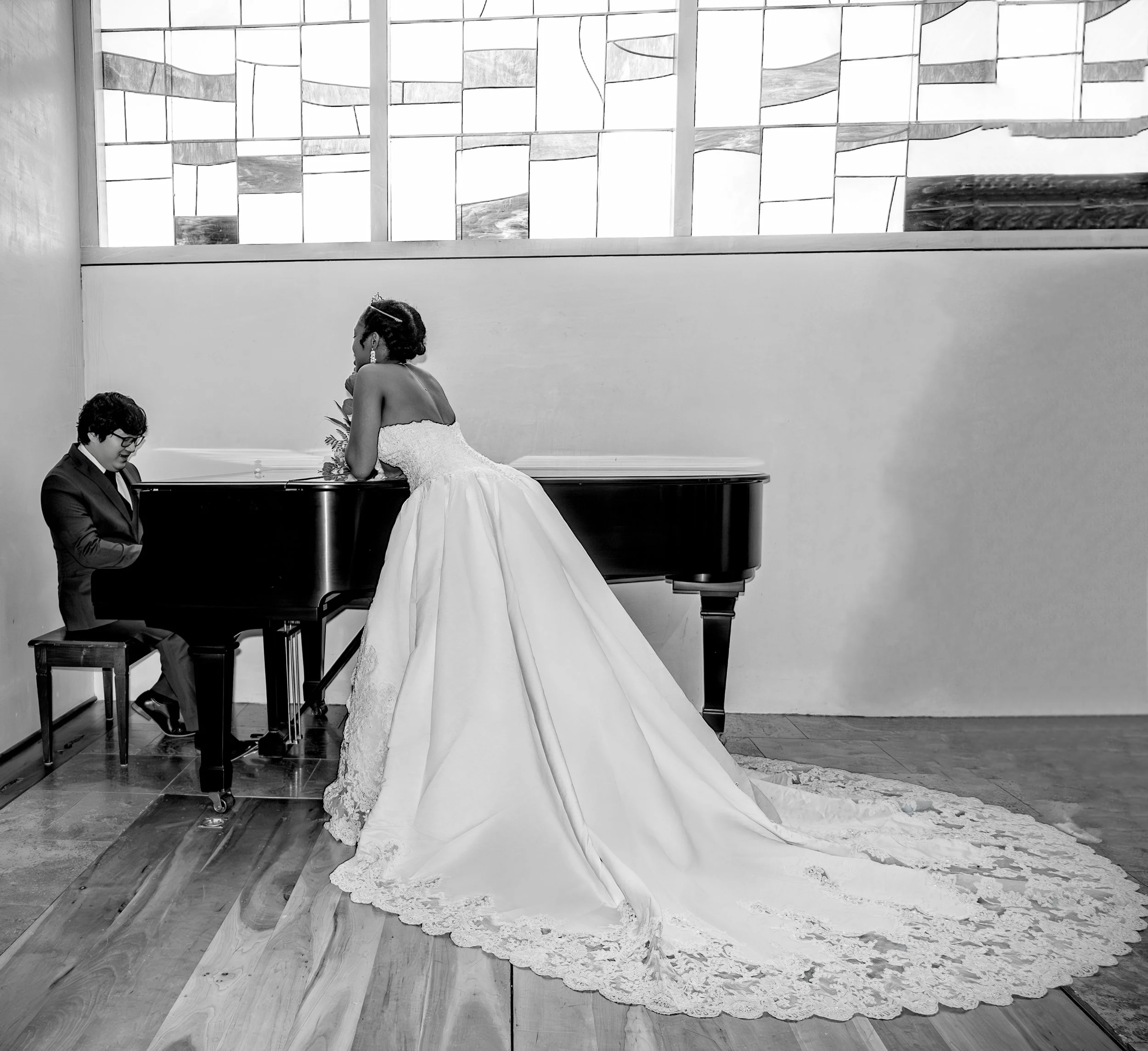 A woman in a wedding dress leaning on a grand piano while a man in a suit sits at the piano, both in a room with stained glass windows. wedding photography dfw photography