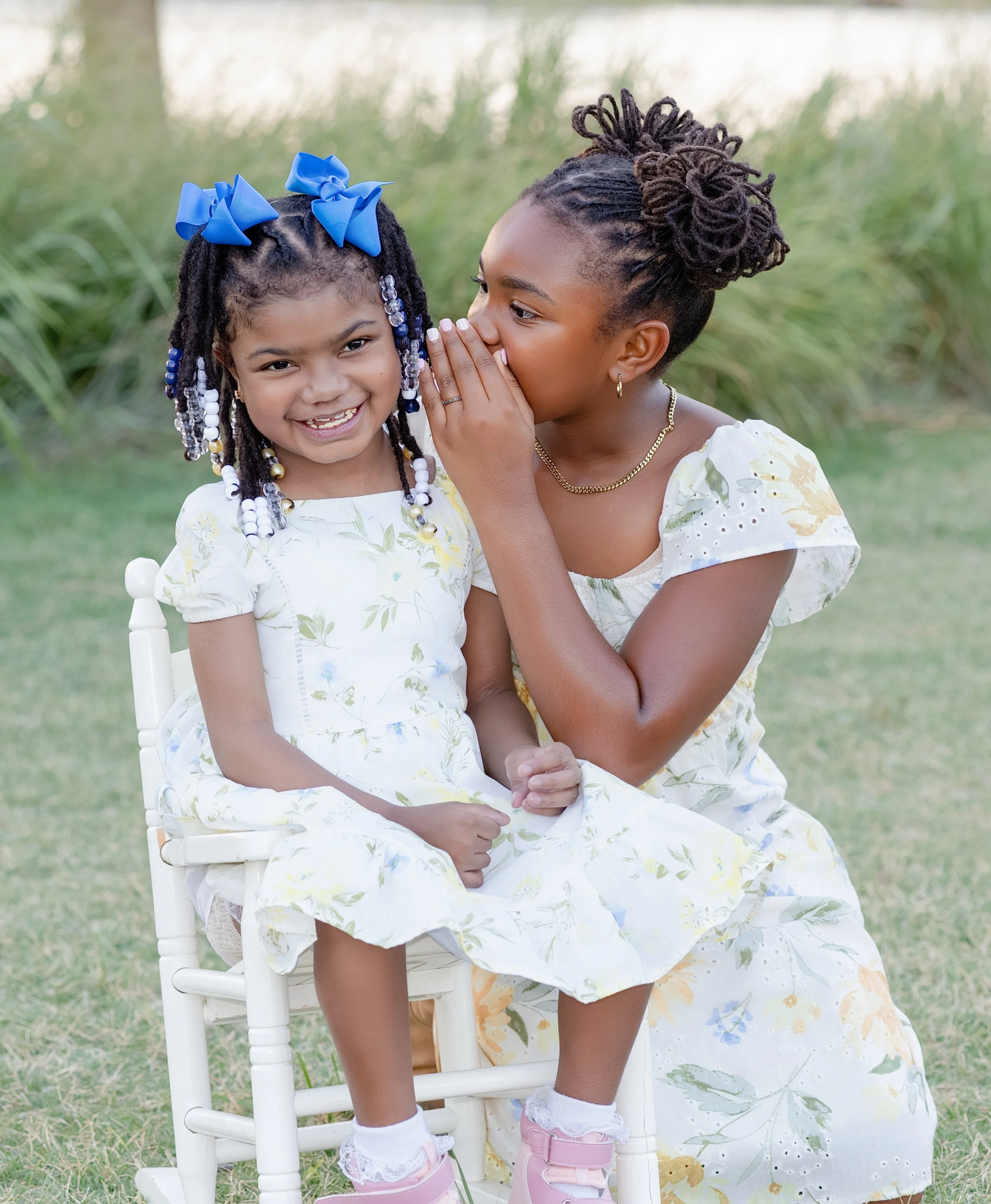 A child whispers a secret to a young girl sitting on a white wooden chair outdoors. The girl has beads in her hair and is smiling. Both girls are wearing floral dresses, and the background is green grass.