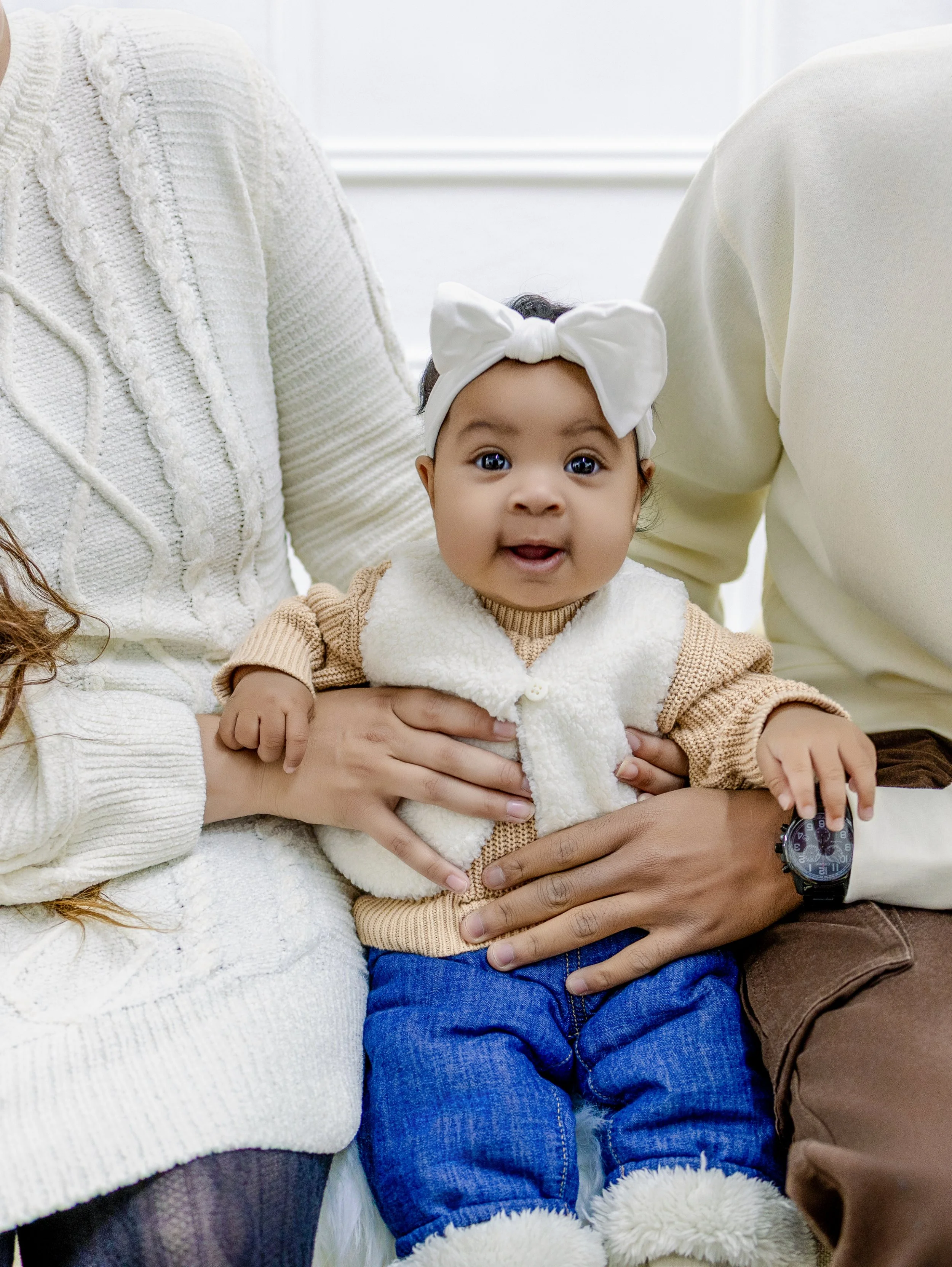 Close-up of a smiling toddler girl with dark hair and big eyes, wearing a white headband with a bow and a cozy beige vest, sitting on someone's lap.