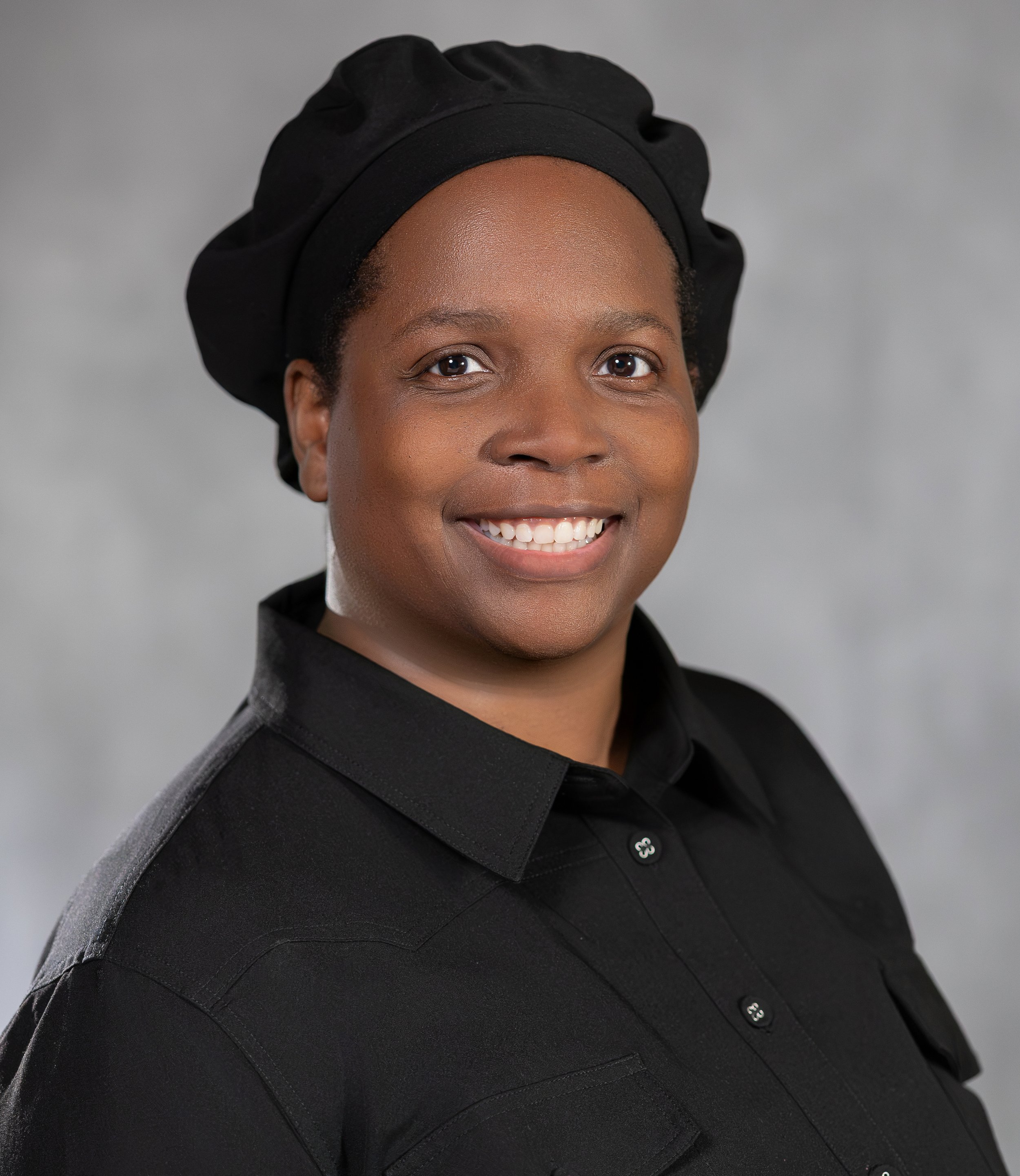 Close-up of a smiling woman wearing a MGT's hat and MGT's coat, against a plain gray background.