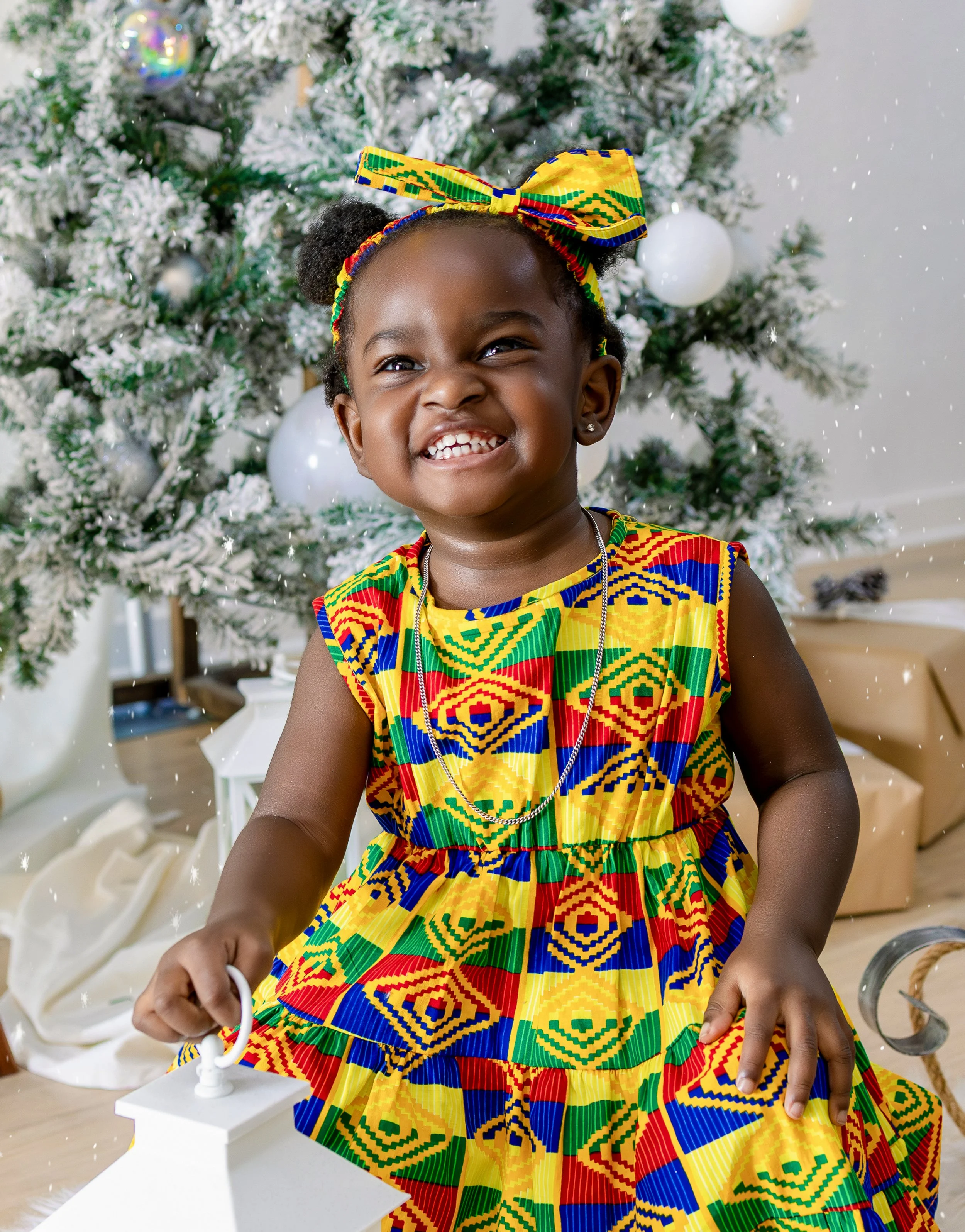 Young girl smiling in colorful African print dress and matching headband in front of a snow-flocked Christmas tree decorated with white ornaments.