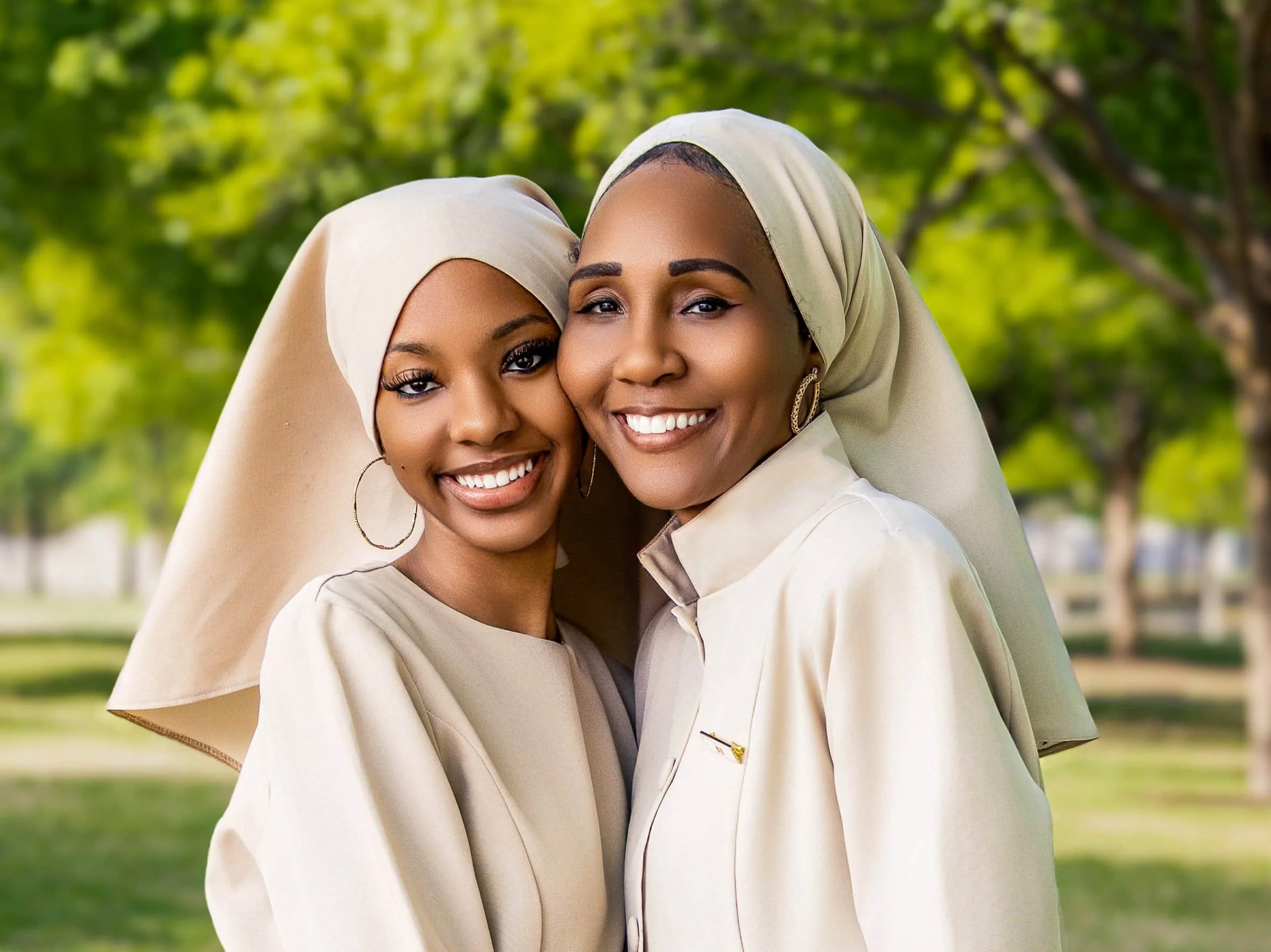 Two women wearing cream-colored outfits and hijabs, smiling outdoors with green trees in the background. DFW Photographer - Family Photographer
