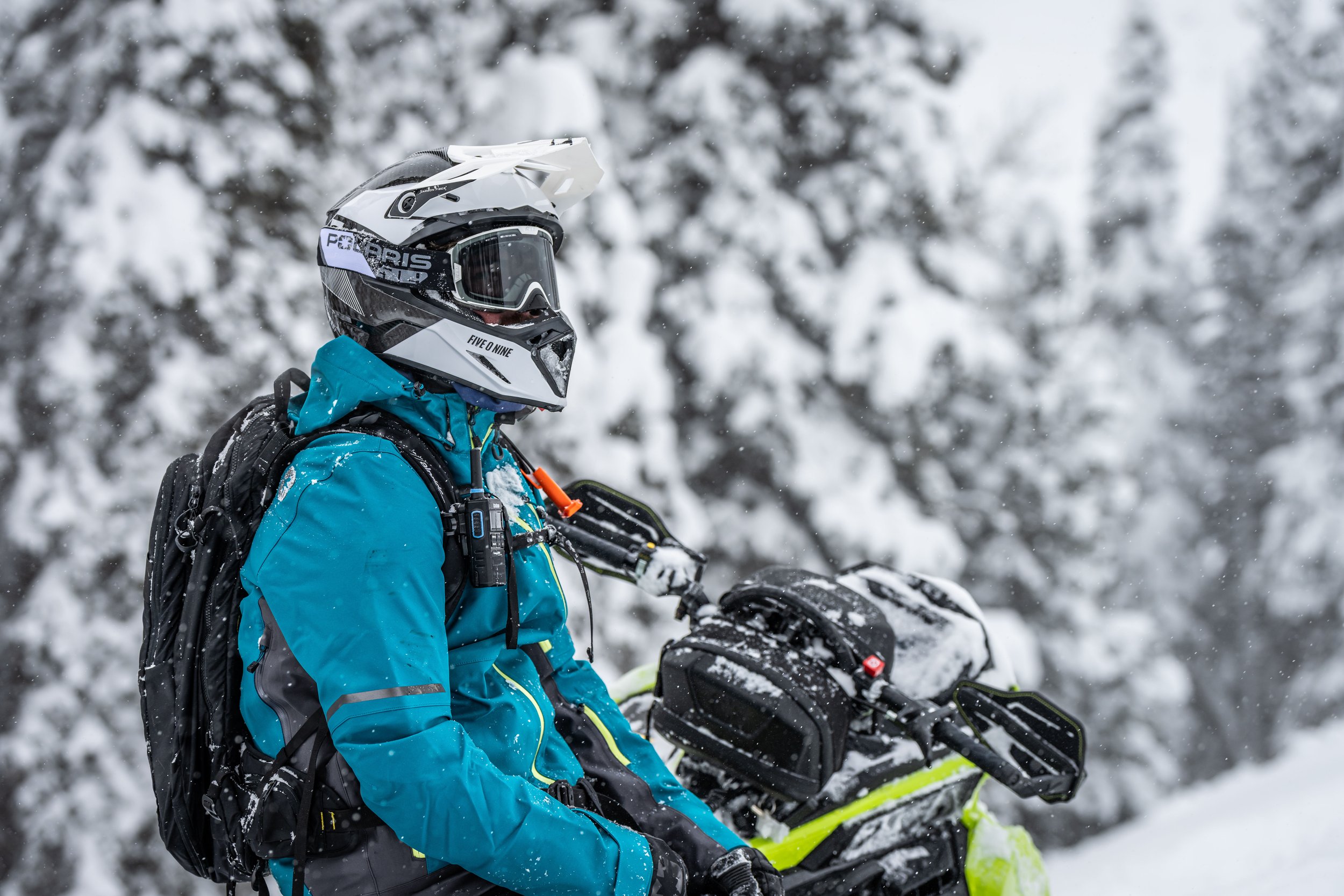 Person in a blue winter jacket with a black backpack and a white snow helmet riding a snowmobile in a snowy forest.