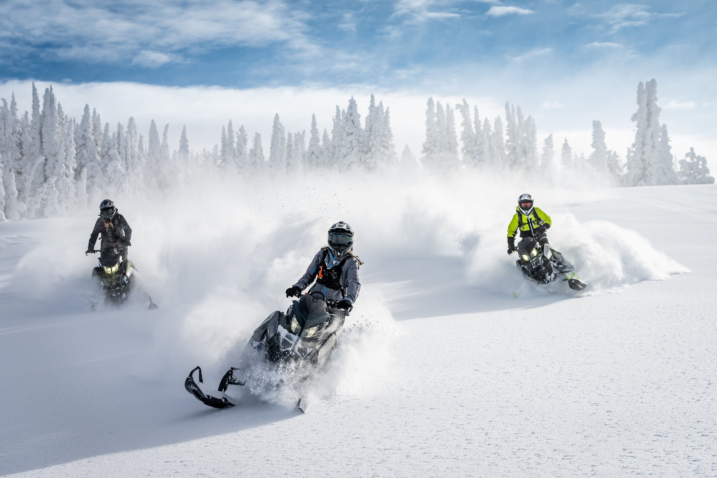 Three people riding snowmobiles across a snowy landscape with snow-covered trees and a partly cloudy sky.