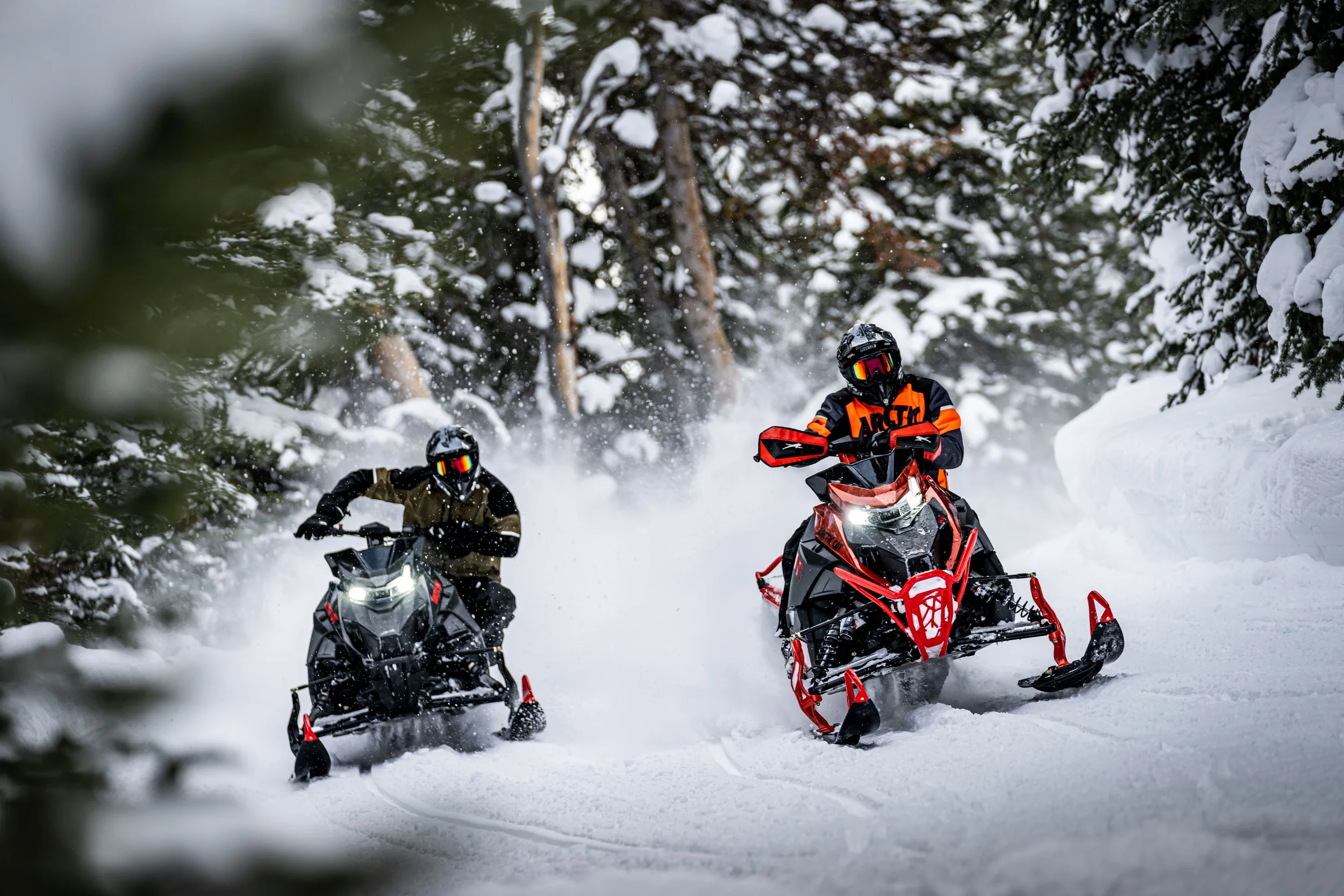 Two people riding snowmobiles through a snowy forested trail with snow-covered trees around them.