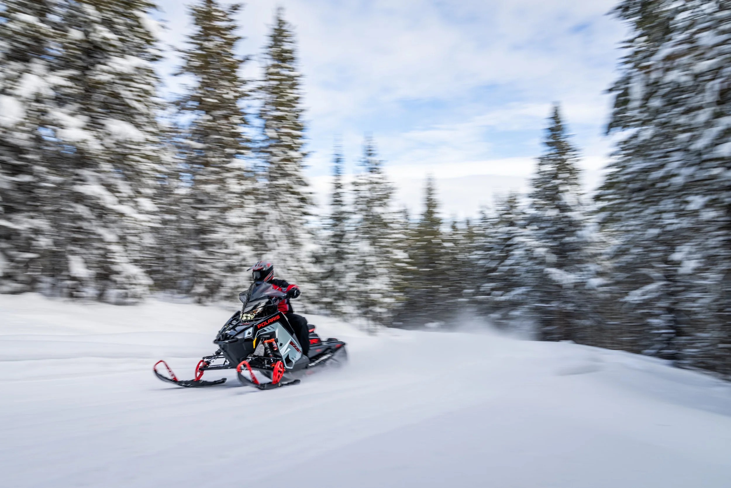 A person riding a Polaris snowmobile through a snowy forest with snow-covered trees and a partly cloudy sky.