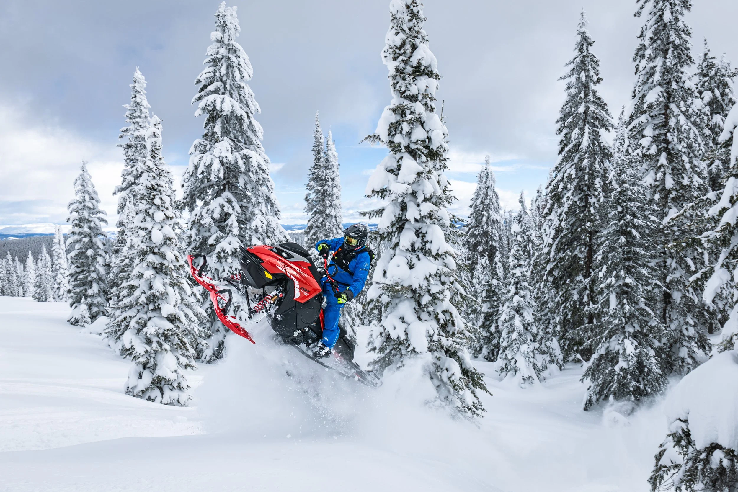 A person riding a red snowmobile through a snowy forest with tall, snow-covered evergreen trees.