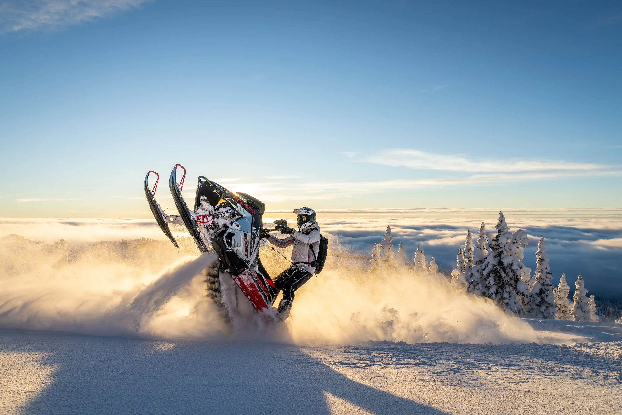 Person riding a snowmobile through snowy terrain at sunset, with snow-covered trees and a clear blue sky in the background.