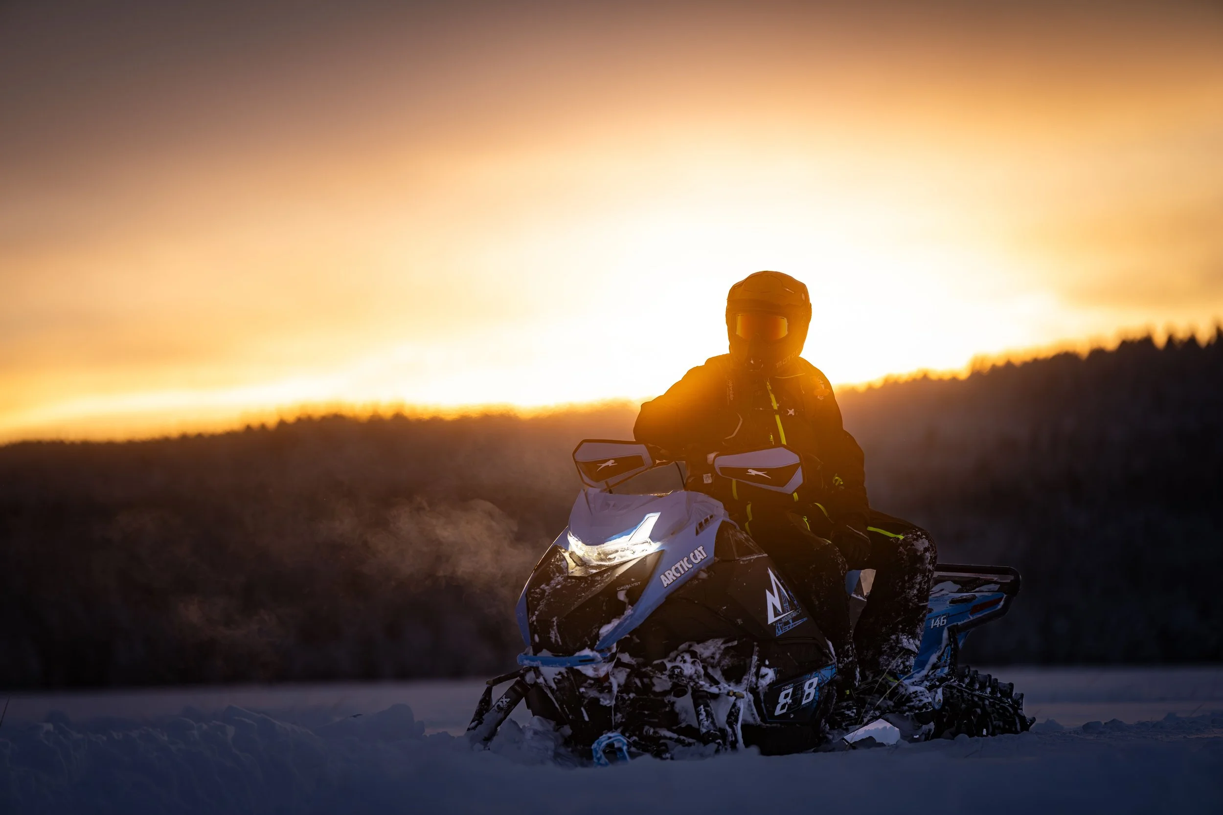 Person riding a snowmobile on snowy terrain at sunset or sunrise, wearing a helmet and winter gear, with a sunset sky in the background.
