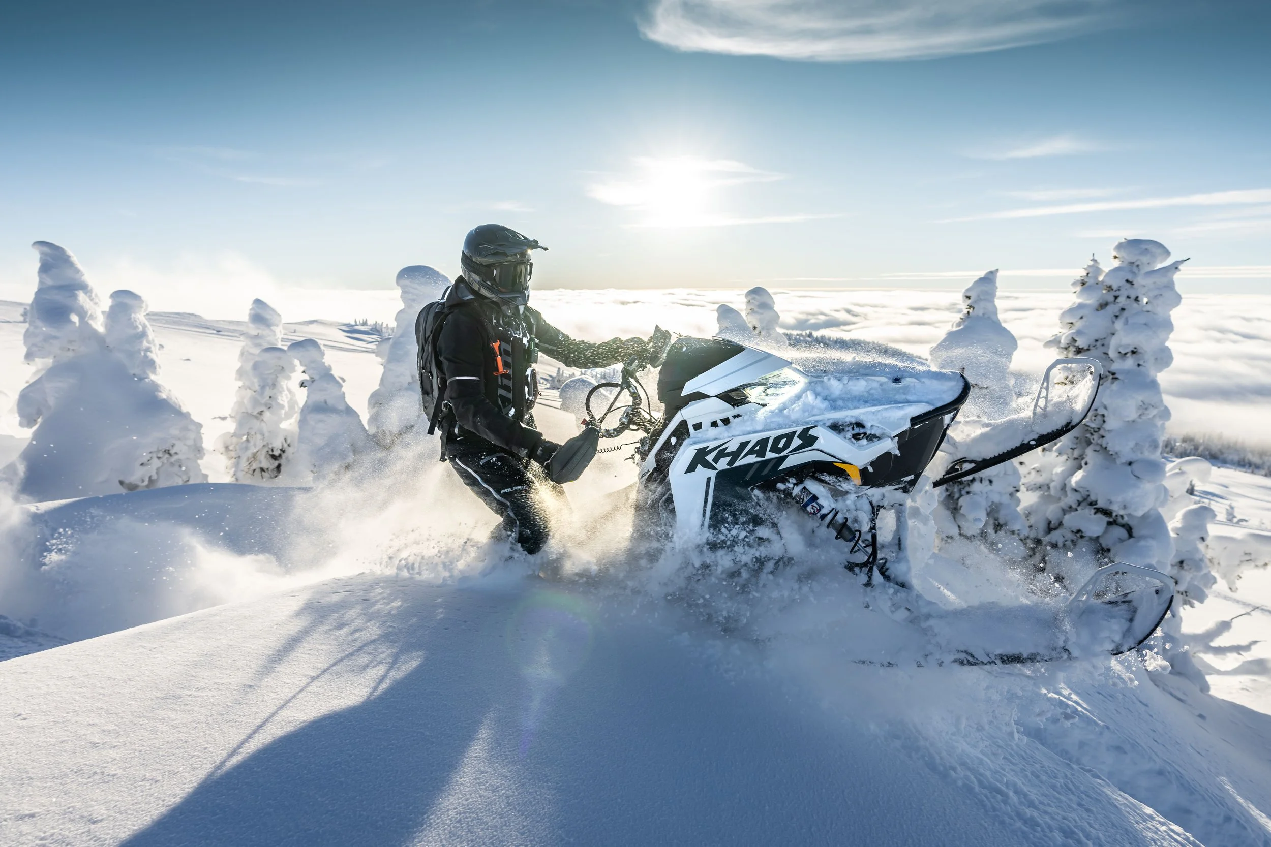 A person in black winter gear and a helmet riding a white snowmobile labeled 'KHAOS' through deep snow in a snowy landscape with snow-covered trees and a bright sun in the sky.