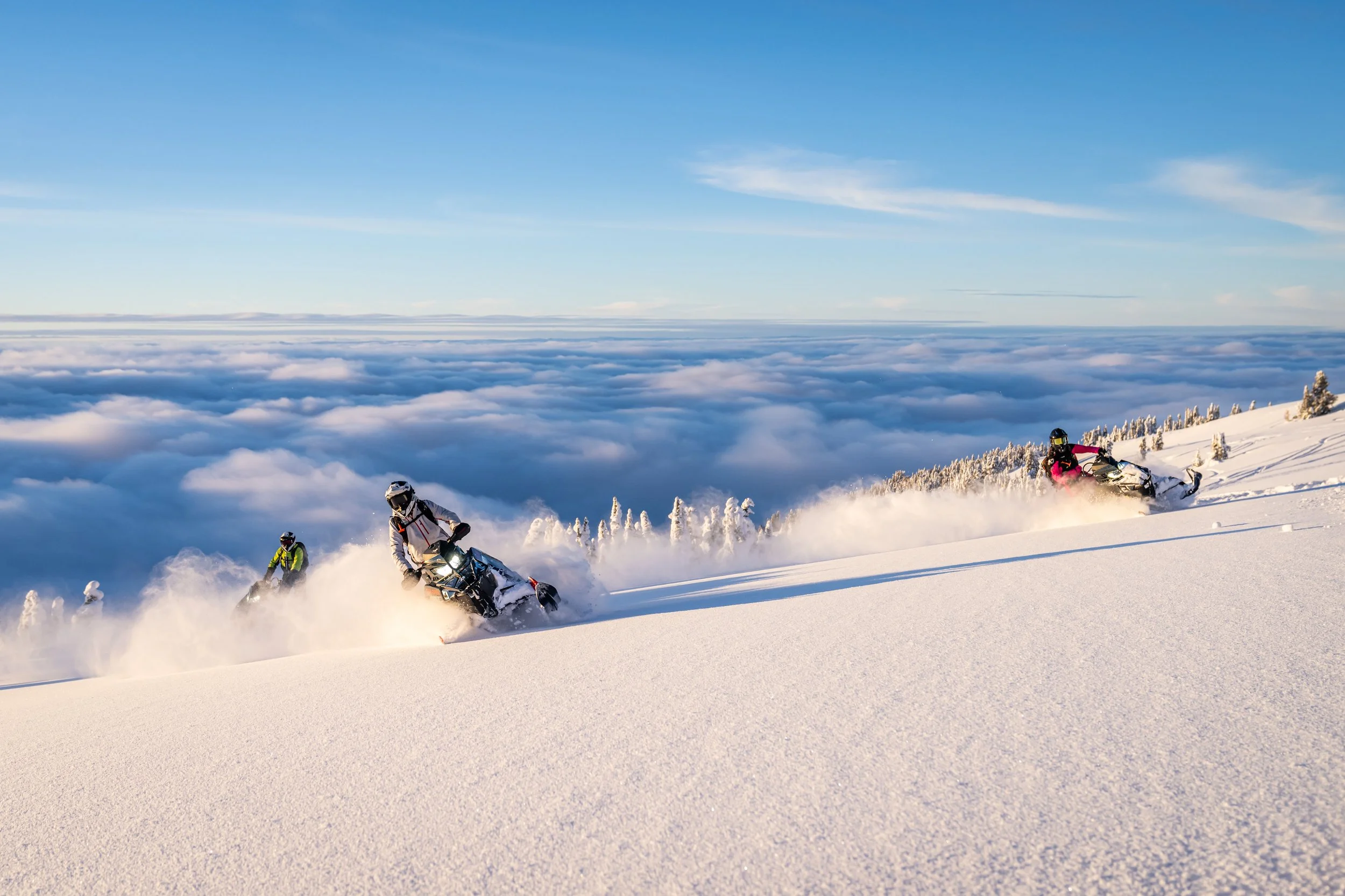 Three snowmobile riders racing down a snow-covered mountain slope under a cloudy sky.