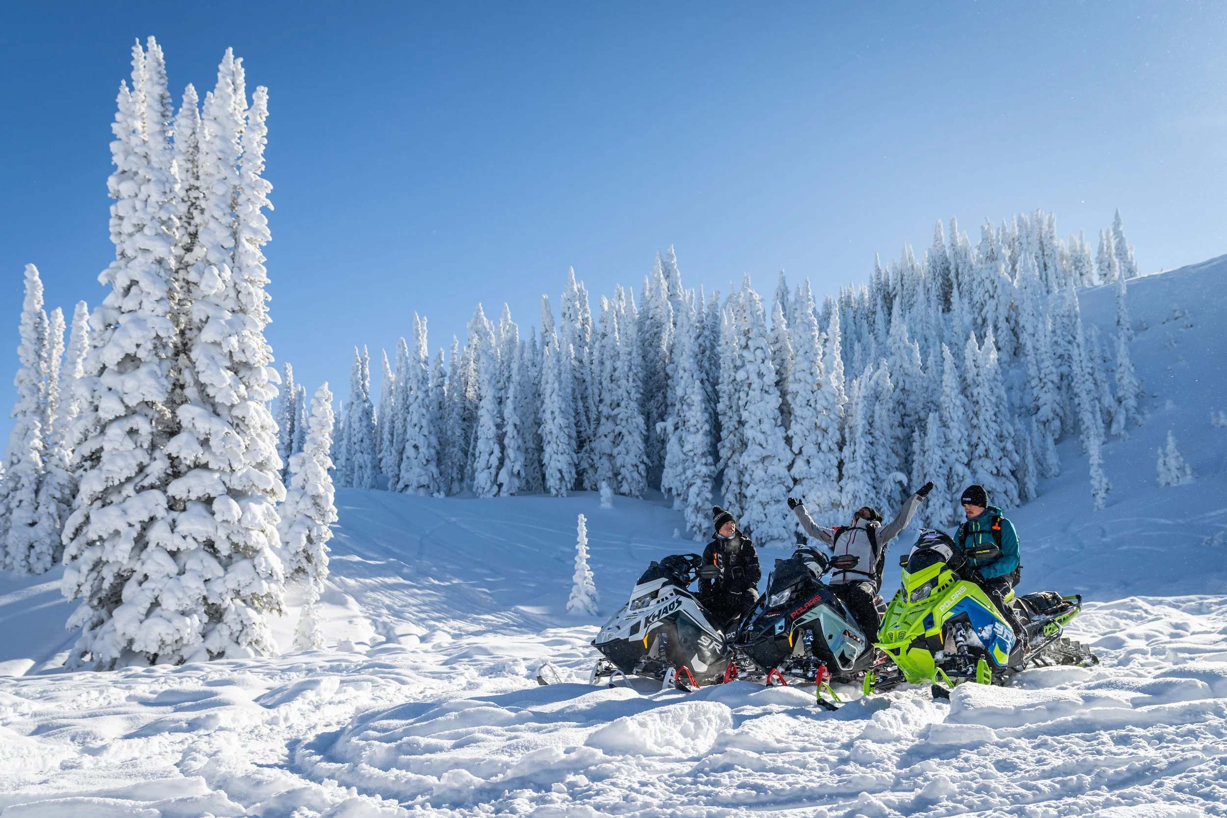 Three people in winter gear sitting on snowmobiles in a snowy, forested landscape with snow-covered trees and a clear blue sky.