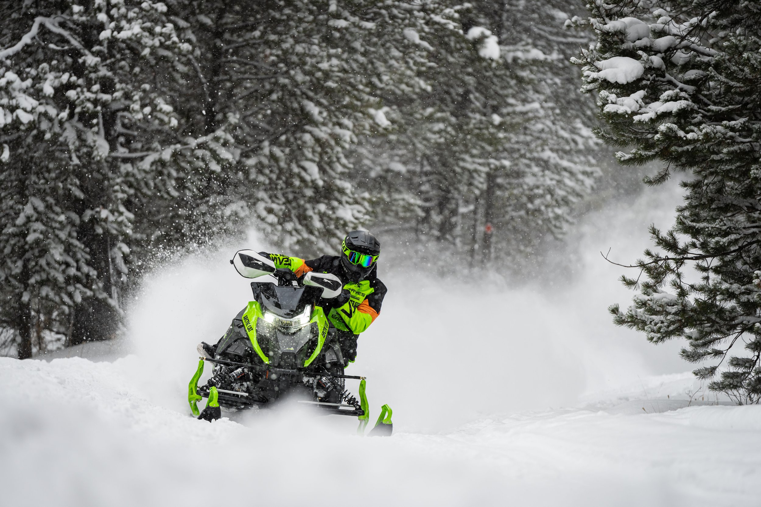 A person riding a snowmobile through a snowy forest, wearing a neon green and black suit with a black helmet and reflective goggles.