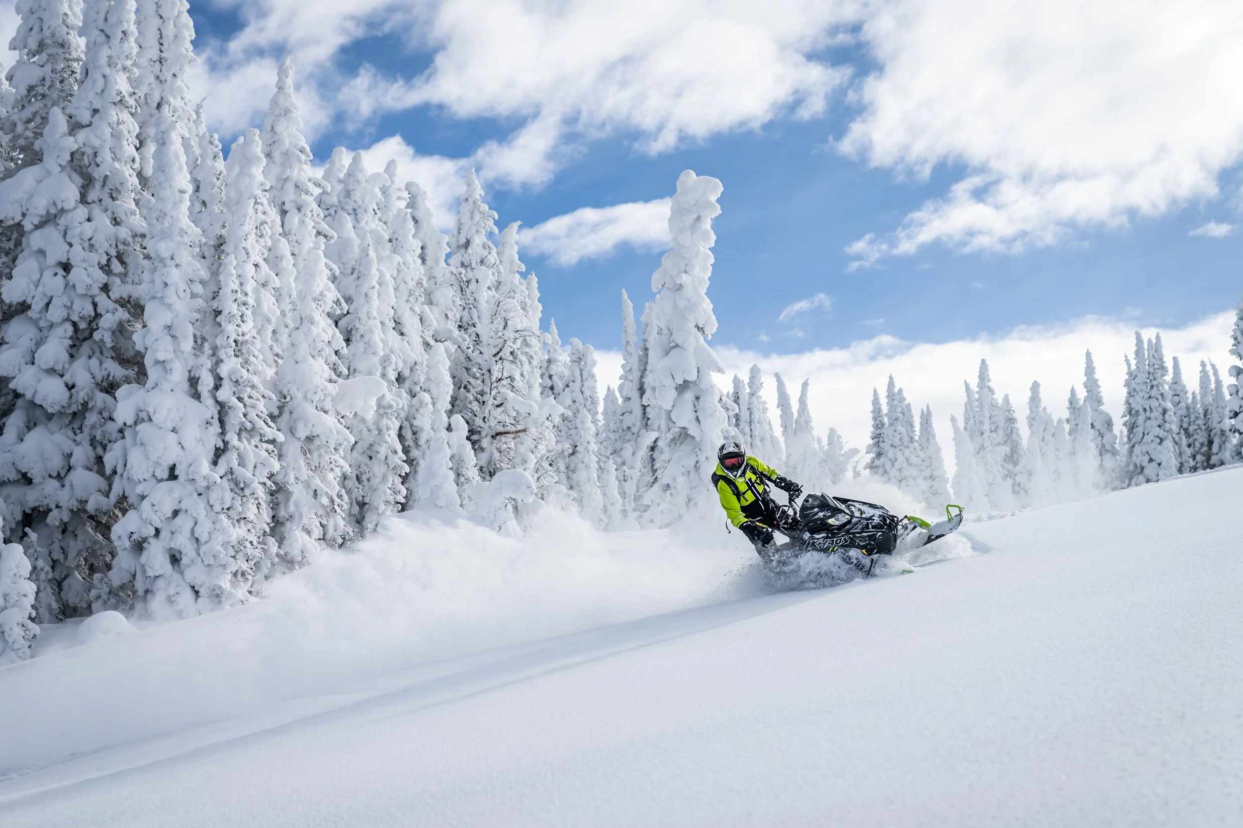 A person riding a snowmobile through a snowy winter landscape with snow-covered trees and a partly cloudy sky.