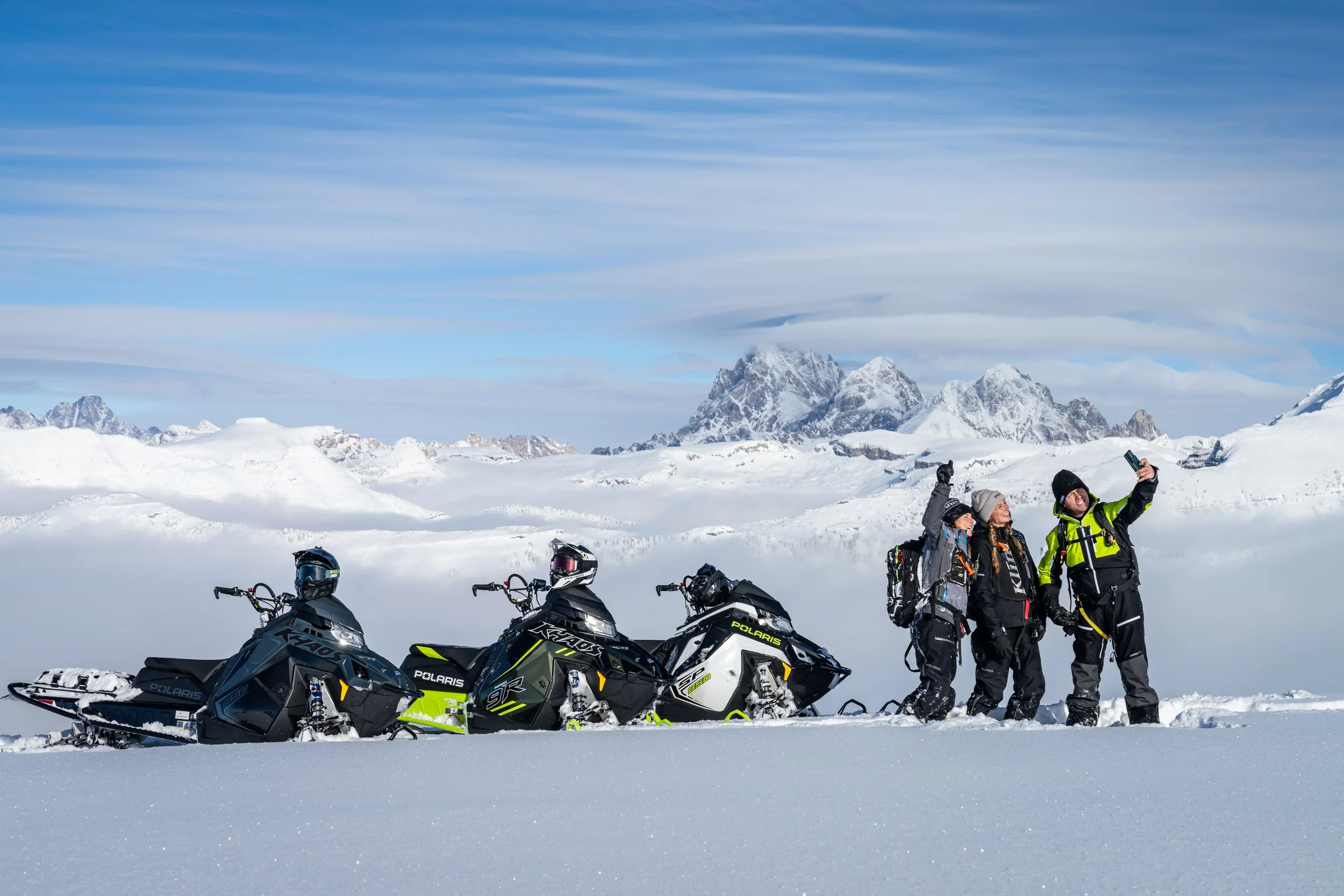 Four people in winter gear taking a selfie in a snowy landscape with all-terrain snowmobiles and snow-covered mountains in the background.