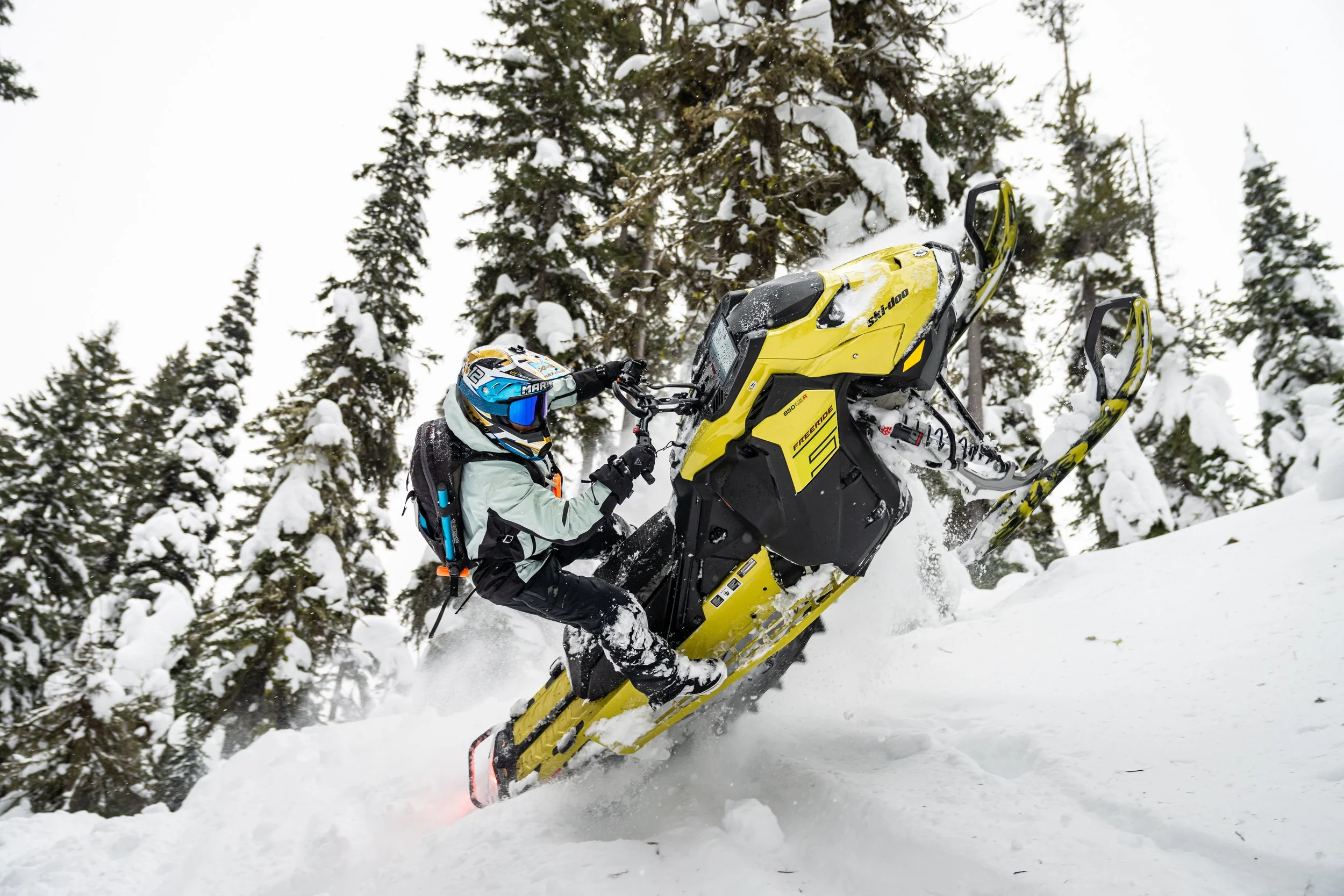 A snowmobile rider in winter gear riding a yellow snowmobile through snowy terrain surrounded by snow-covered trees.