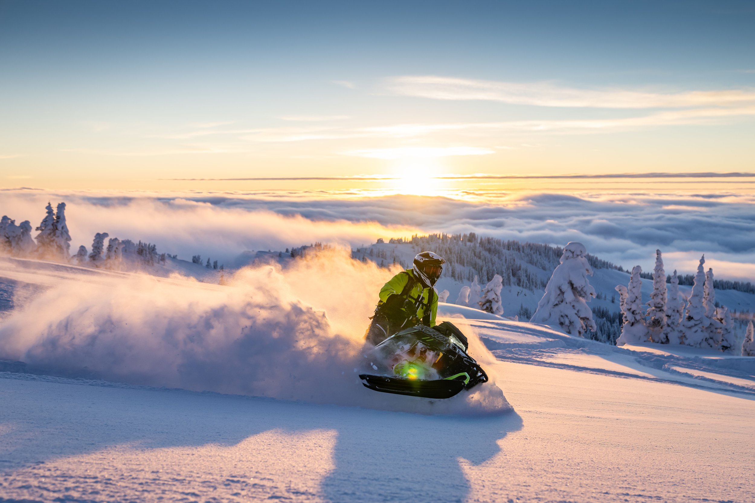 A person riding a snowmobile in snowy mountains during sunset with snow-covered trees in the background.