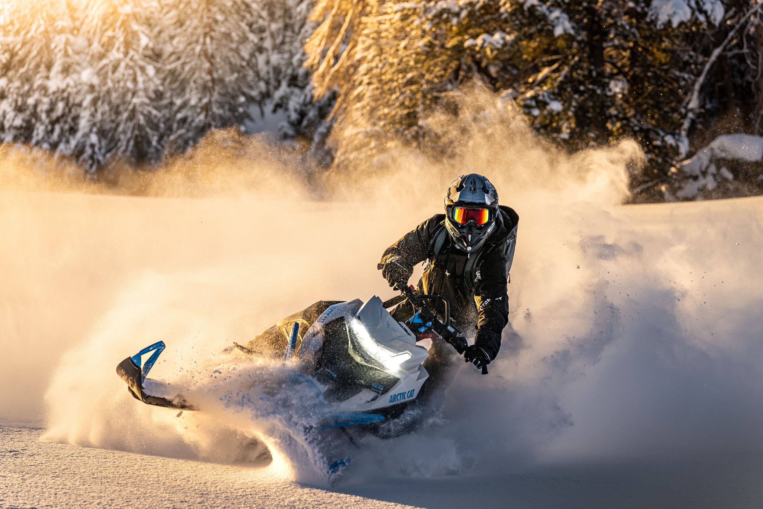 A person riding an Arctic Cat snowmobile through snow in a winter landscape with snow-covered trees in the background, during sunset or sunrise.